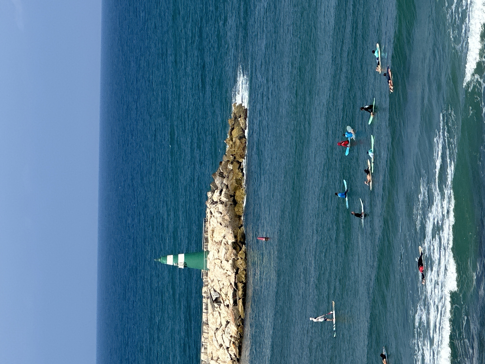 The image depicts a scene at the Tel Aviv port, Israel, with a lighthouse at the end of a rock jetty. The sea is calm and blue, with small waves breaking near the shore. Several people are engaged in water sports. In the foreground, on the left, a person stands on a paddleboard. Further out in the water, a group of individuals are on paddleboards and surfboards. One person is lying prone on a surfboard in the whitewater. In the middle ground, there is a red buoy. The lighthouse is green and white, situated on a rocky embankment. The sky is clear and blue.