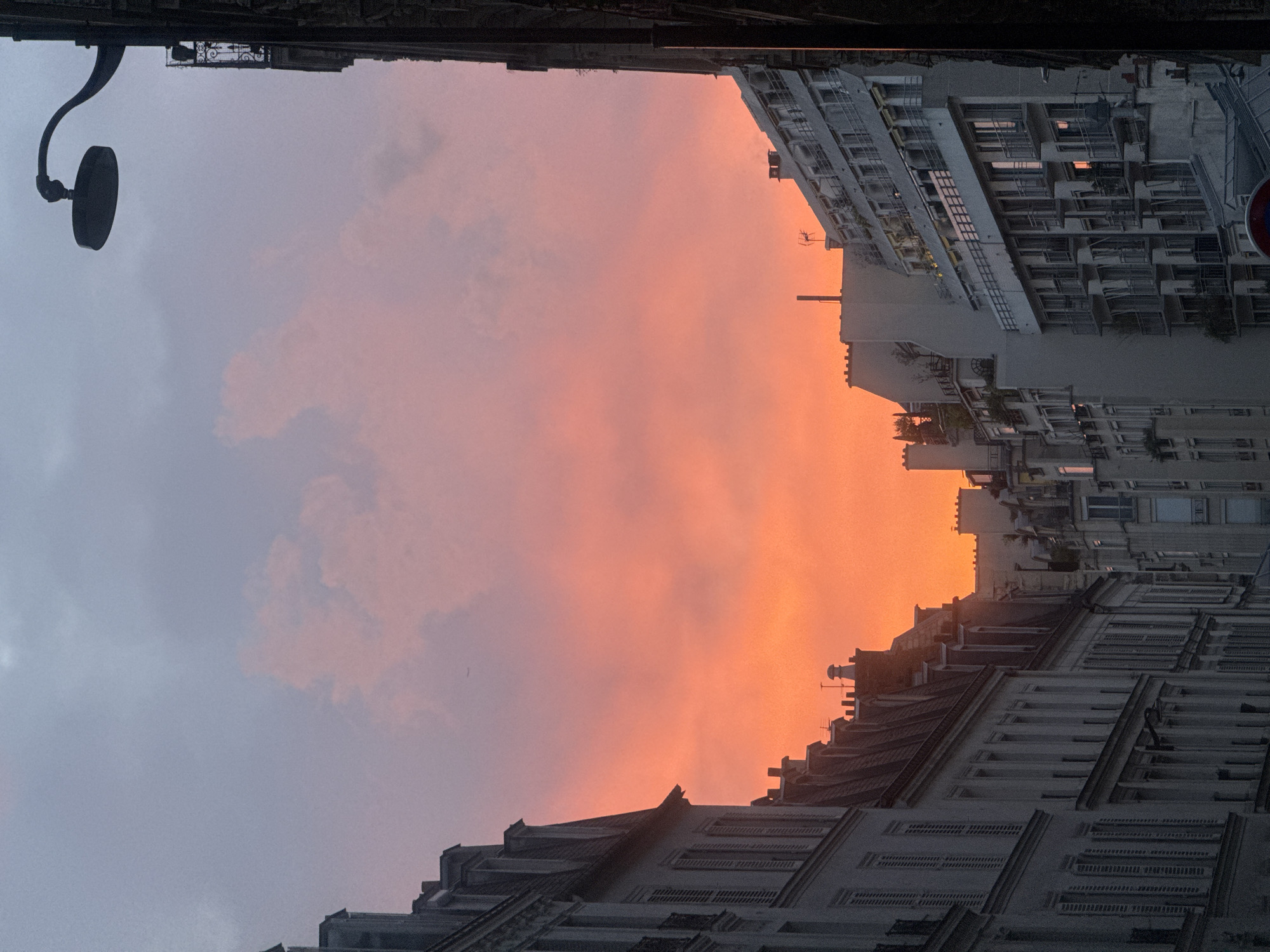 The image depicts a view of Paris, France, at dusk or dawn. The sky is colored with hues of orange and pink near the horizon, indicating either a sunrise or sunset. The buildings on either side of the frame are typical of Parisian architecture, with multiple windows and detailed facades. The buildings are likely residential or mixed-use structures, common in the city center. The image is oriented upside down, as if the viewer is looking upwards from between buildings. A street lamp is visible on the left side of the frame.