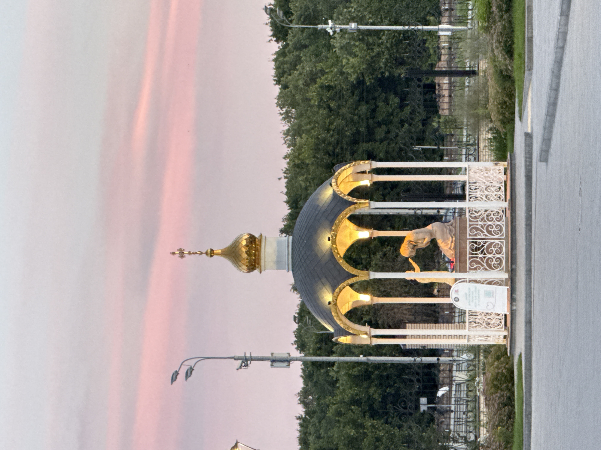 The image shows a close-up view of a golden dome, possibly part of a church or an ornate structure, with a white decorative railing surrounding it. The dome has an onion shape and is adorned with gold accents. Several lights illuminate the underside of the dome structure, creating a warm glow. A statue of a person, seemingly bending or reaching, is visible within the fenced area. The background consists of lush green trees and a pale sky with soft pink hues, suggesting either sunrise or sunset. In the foreground, a paved walkway runs along the right side of the image. A streetlamp is visible to the left of the dome structure, and a white sign with some text and icons is placed near the statue. The scene appears to be set in an outdoor public space. The image is oriented vertically. The time of day is likely dawn or dusk due to the soft light and sky color. There are no explicit indicators of specific weather conditions, but the sky suggests fair weather.