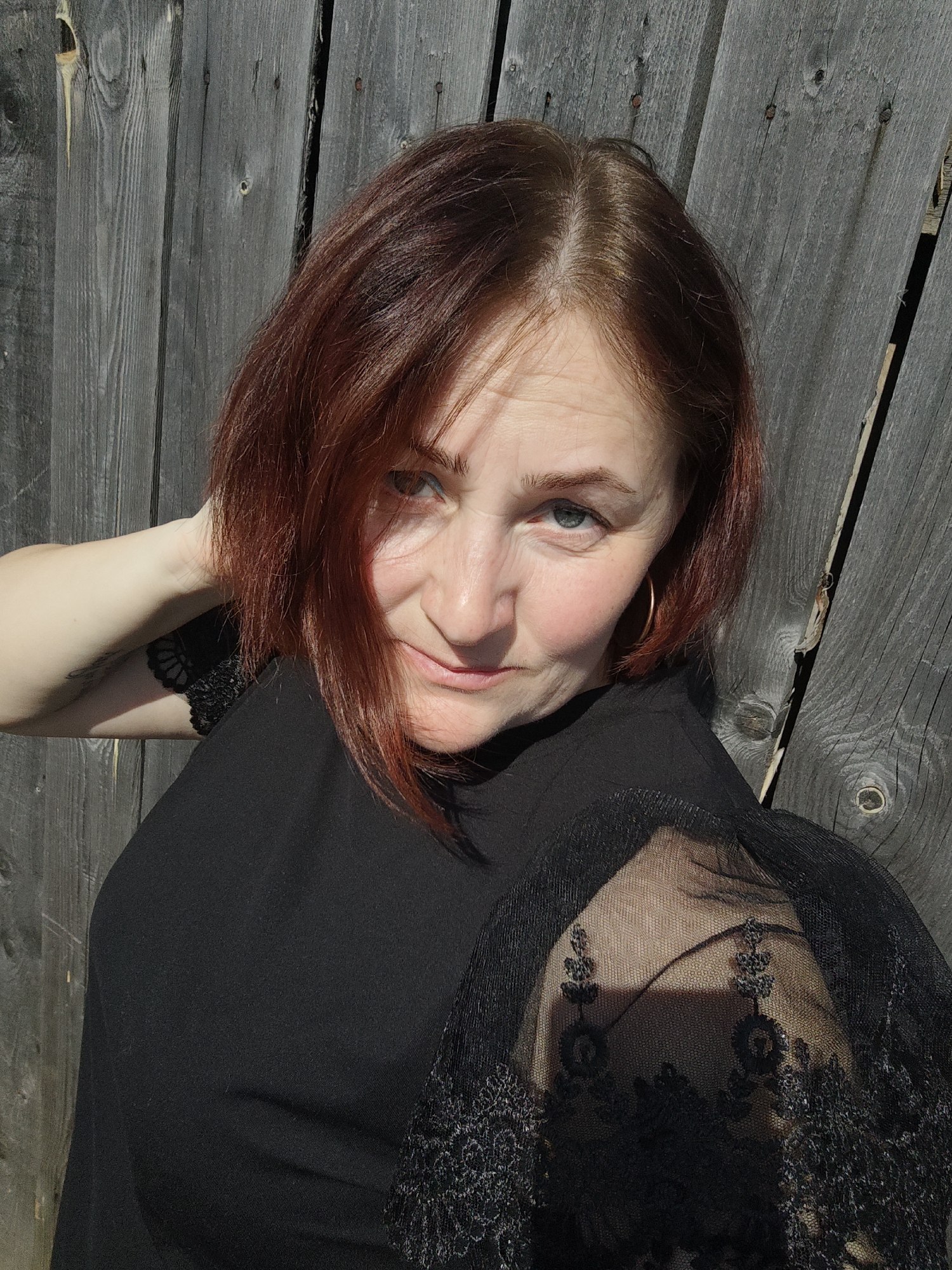 A woman poses in front of a gray wooden fence. She has short, reddish-brown hair and blue eyes. She is wearing a black top with lace sleeves. Her left arm is raised behind her head. She appears to be in an outdoor location, possibly in the city of Фоки, Russia.