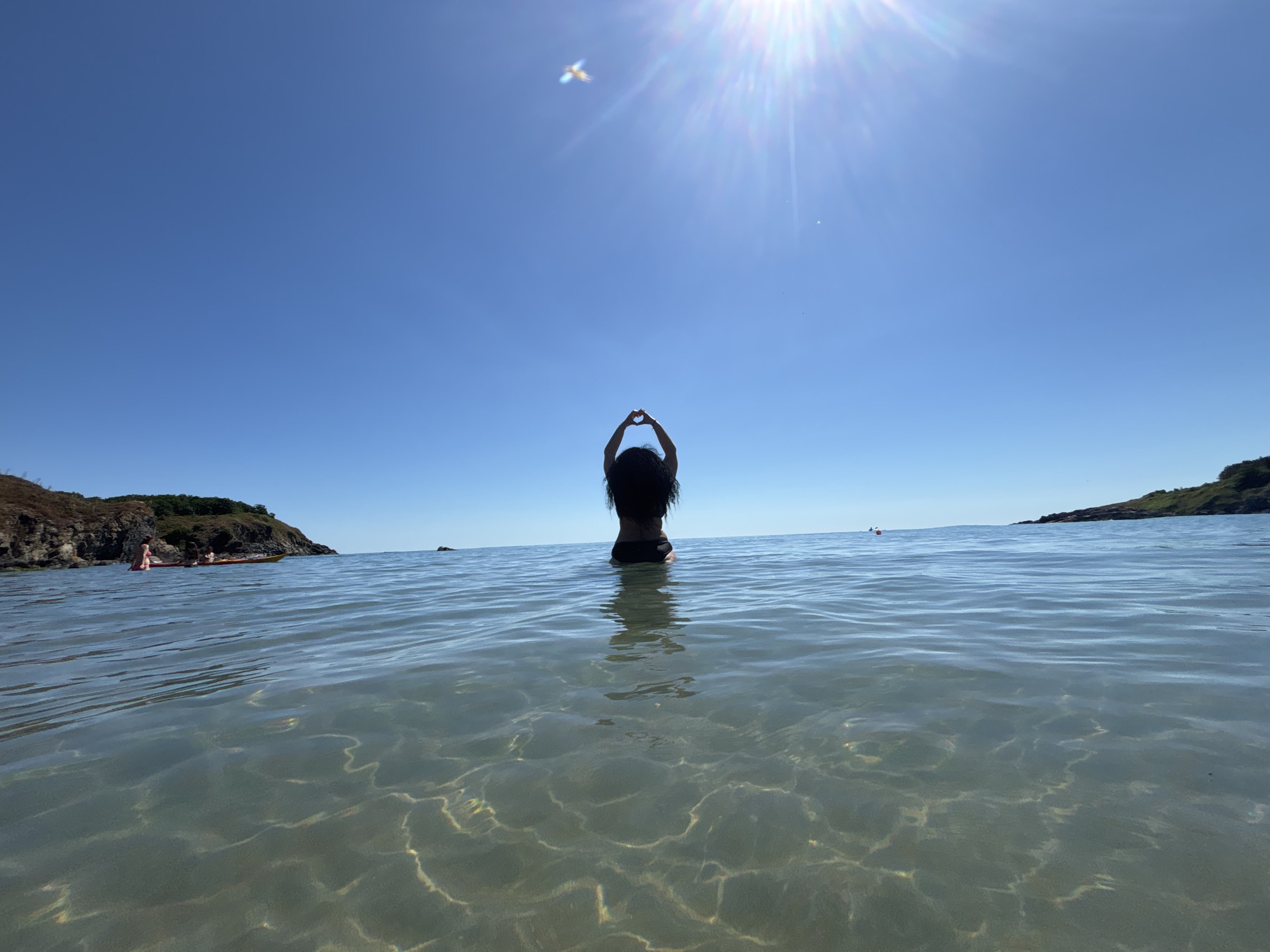 A low-angle, wide-shot photograph captures a person from behind, standing in shallow, clear ocean water with a sandy bottom visible. The person, with long dark hair and wearing dark swimwear, is facing away from the camera and has their arms raised to form a heart shape above their head. The sun is shining brightly in the upper right portion of the frame, creating lens flare and casting shimmering patterns on the water's surface. In the distance, the sea stretches to the horizon under a clear blue sky. On the left side of the image, a rocky, vegetated coastline is visible, with two people in kayaks near the shore. On the right side, another rocky coastline is visible in the distance. The overall scene depicts a serene and sunny day at the beach in Rezovo, Bulgaria.