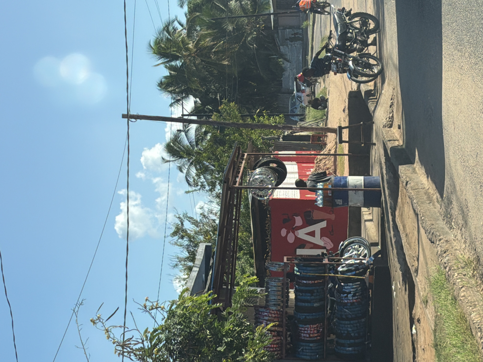 The image captures a vibrant street scene under a bright, sunny sky with a few scattered clouds. The primary focus is a roadside shop specializing in motorcycle parts, visible on the left side of the frame. This shop features metal racks heavily laden with stacks of colorful motorcycle tires and inner tubes. A prominent red sign on the shop's facade displays the partial text "IA". A blue and white barrel is positioned near the front of the shop.
Several people are present in the scene. One individual stands near the blue barrel, while another person is seated on a motorcycle further down the dirt shoulder, appearing to be working on it. More motorcycles and people are visible in the background, indicating a bustling environment. One person in an orange vest is seen riding a motorcycle further down the street.
The setting is characterized by abundant green foliage, including tall palm trees, suggesting a tropical or subtropical location. Power lines are visible overhead, strung between utility poles. A paved road runs along the right side of the image, bordered by a concrete curb and an unpaved, dirt or gravel shoulder where the motorcycles are parked. The strong sunlight casts sharp shadows, confirming it is daytime. The scene conveys the activity of a local motorcycle repair or parts sales business in a lively, informal setting.