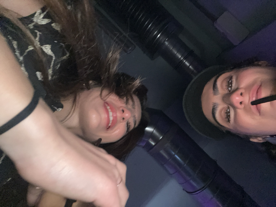 Two smiling women in a dimly lit area are captured from a low angle. The woman on the left has dark hair, a floral patterned top, and is holding a phone or camera with a visible hand. The woman on the right has a hat and is drinking from a straw. Black pipes or ventilation ducts are visible in the background. The lighting is subdued, giving a club-like atmosphere.