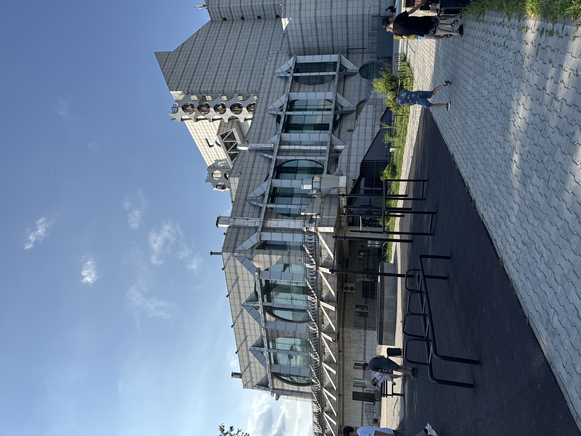 The image shows an outdoor scene in Blagoveshchensk, Russia. In the background, a large, modern building with a gray, tiled facade is visible. The building has a distinctive design with geometric shapes, circular windows, and angular protrusions. In the foreground, there is an outdoor fitness area with black rubber flooring and metal exercise equipment. A paved walkway made of gray interlocking bricks runs alongside the fitness area. Several people are present in the scene, including individuals walking, sitting on a bench, and a child running. The sky is blue with scattered white clouds. The overall environment suggests a public space designed for recreation and leisure.