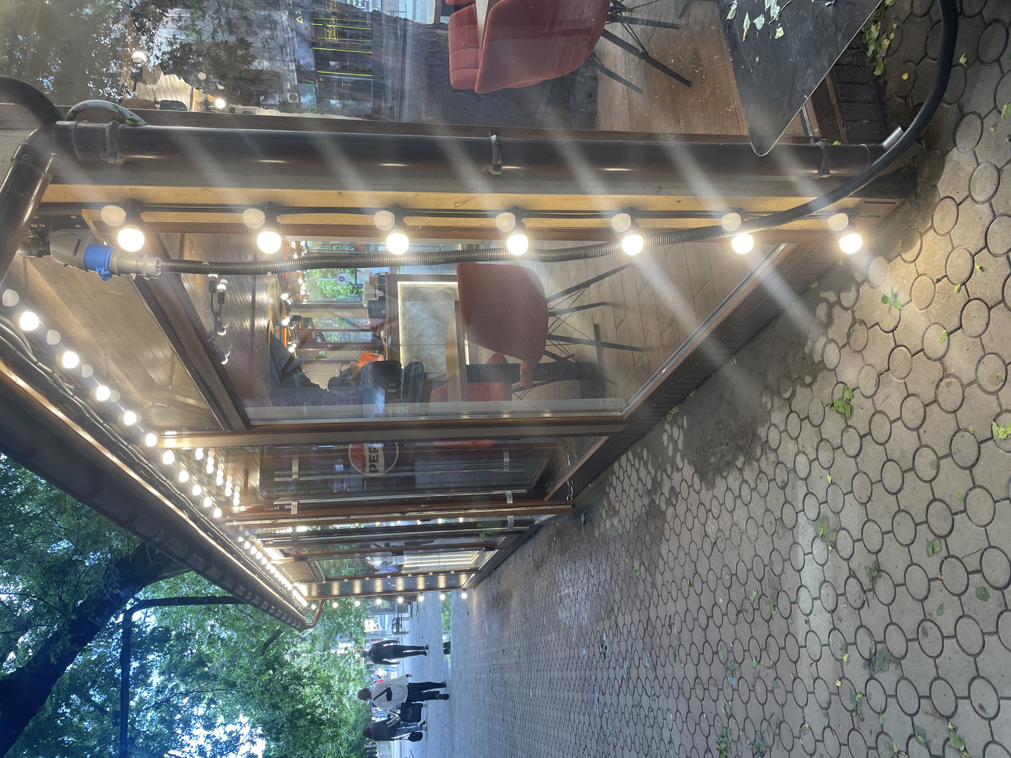A street view shows the exterior of a cafe in Mykolaiv, Ukraine. The cafe has a series of interconnected enclosed outdoor seating areas with glass walls and string lights illuminating the space. Inside, tables and chairs are visible. The facade of the cafe displays a "PEPSI" logo. A paved sidewalk runs alongside the cafe, and several people are walking on it. A tree with green leaves is visible, suggesting an urban environment.