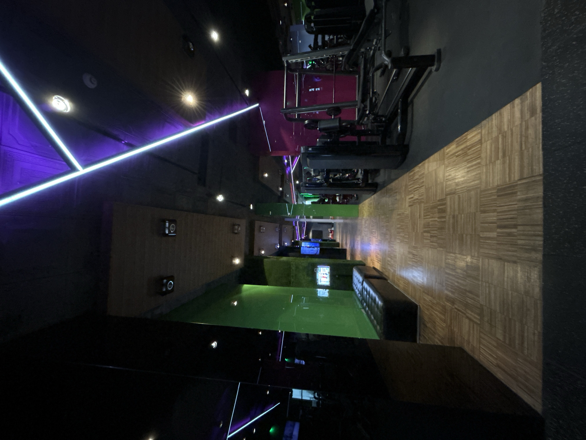 The image depicts the interior of a modern-style room. The walls are covered in a wooden tile pattern. There is a set of gym equipment on the right side of the room. On the left side is a lounge area with green flooring and dark, built-in couches. Above the lounge area, lights create a purple hue in the ceiling, and on the bottom, there are black panels with blue and green lights. The image does not provide enough information to determine the specific location within the city of Plovdiv, Bulgaria.