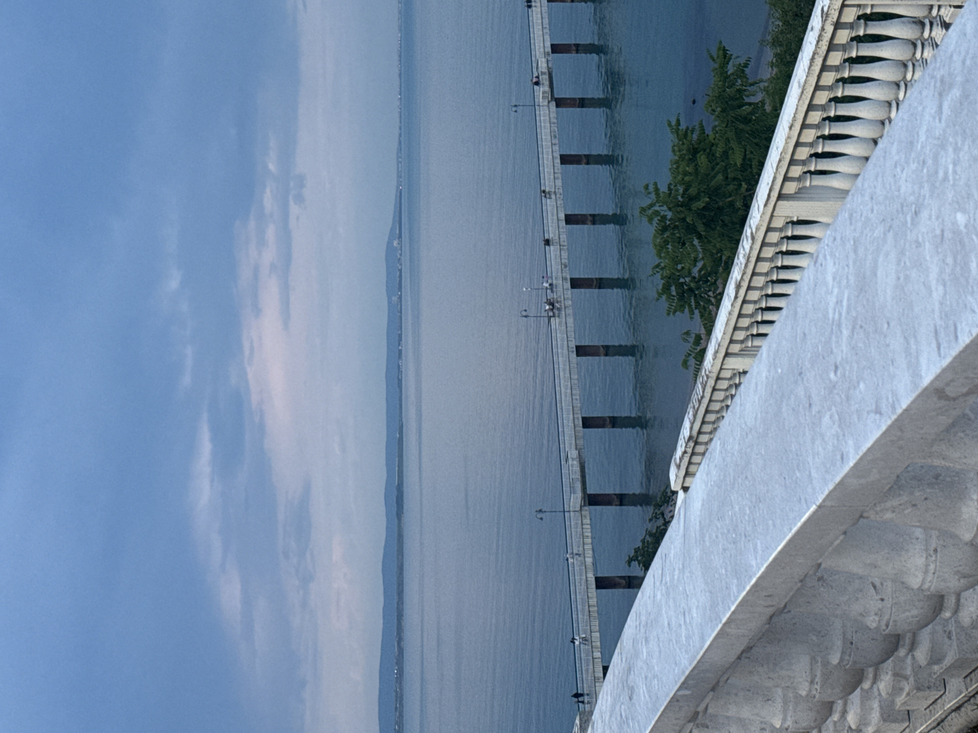 The image captures a tranquil scene, likely at twilight, from a viewpoint along a decorative balustrade. The foreground is dominated by the balustrade, constructed of gray stone with repetitive, rounded balusters. Beyond the balustrade stretches a large body of water, likely the Black Sea, its surface calm and reflecting the muted light. A long pier extends into the water, supported by evenly spaced pilings. Small figures of people are visible along the pier. The sky is partly cloudy, transitioning from a lighter hue near the horizon to a darker blue higher up. In the distance across the water, there is a hint of land. The location is possibly the Sea Garden promenade in Burgas, Bulgaria, due to the architectural style of the balustrade and its location along the coast.