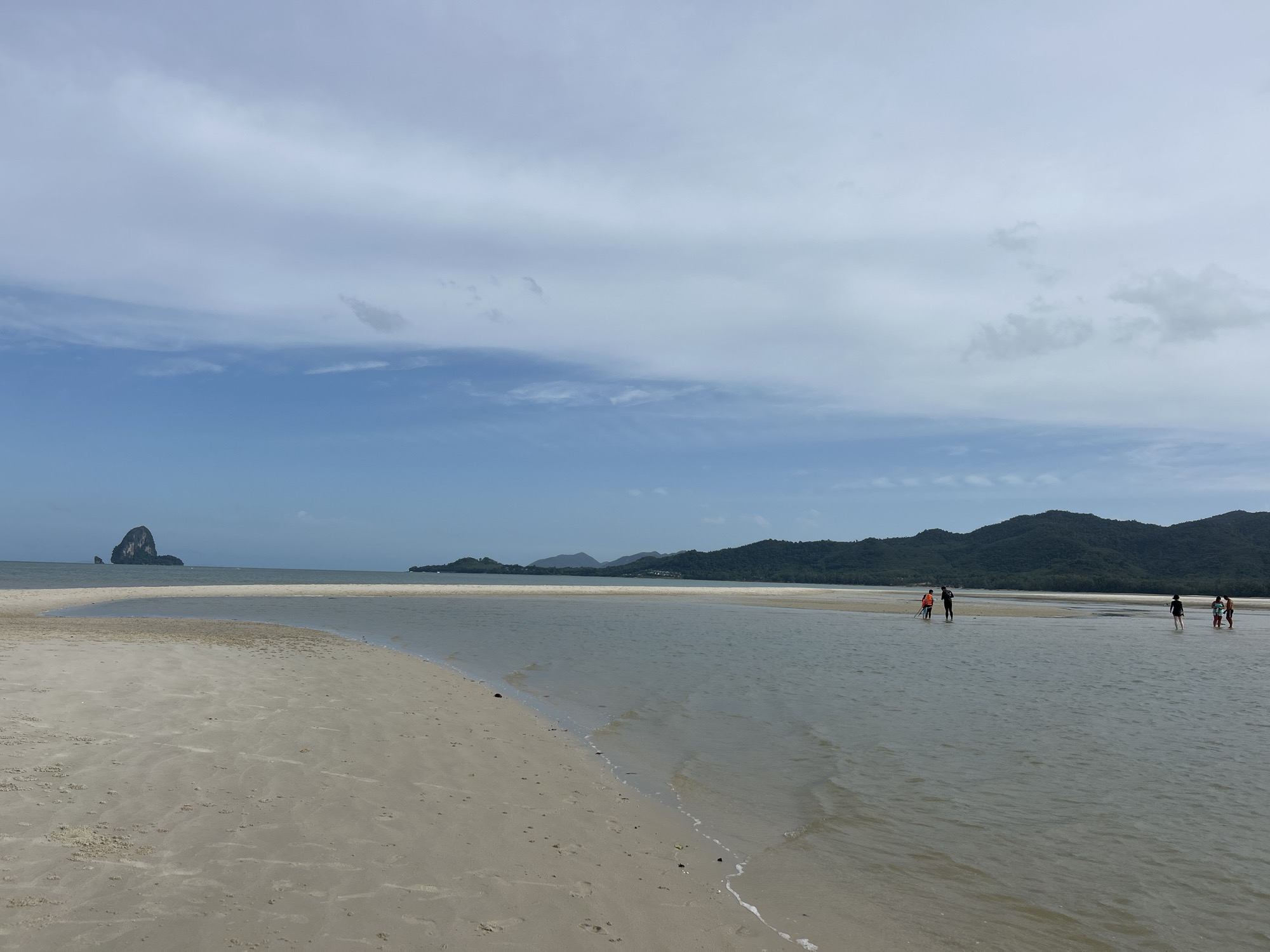 The image captures a serene beach scene in the region of Ko Yao, Thailand. The foreground is dominated by a vast expanse of light-colored sand, showing some subtle textures and footprints, leading to the gentle lapping of shallow water. In the mid-ground, a wide, calm body of water stretches out, with a sandbar visible beneath the surface in places. Further in the distance, a chain of lush green islands and mountains forms the horizon, with a distinct, conical rock formation emerging from the sea to the left. The sky above is a blend of soft blue and diffused white clouds. Several individuals are present in the shallow water, appearing as small figures engaged in leisurely activities. Two people in the mid-ground are walking away from the viewer, while a group of three are further to the right, appearing to stand and converse or observe the surroundings.