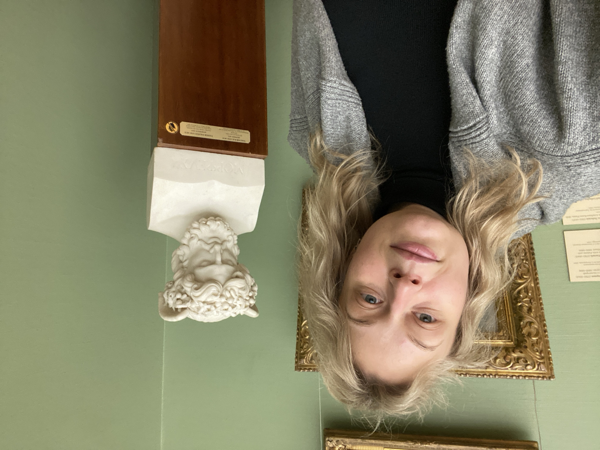 A pale-skinned woman with blonde, shoulder-length hair, wearing a black turtleneck and grey cardigan, poses in front of an off-white bust on a brown wooden plinth in a room with green walls. In the background are two gold-framed artworks and informational plaques on the wall. The location is the State Tretyakov Gallery in Moscow, Russia.