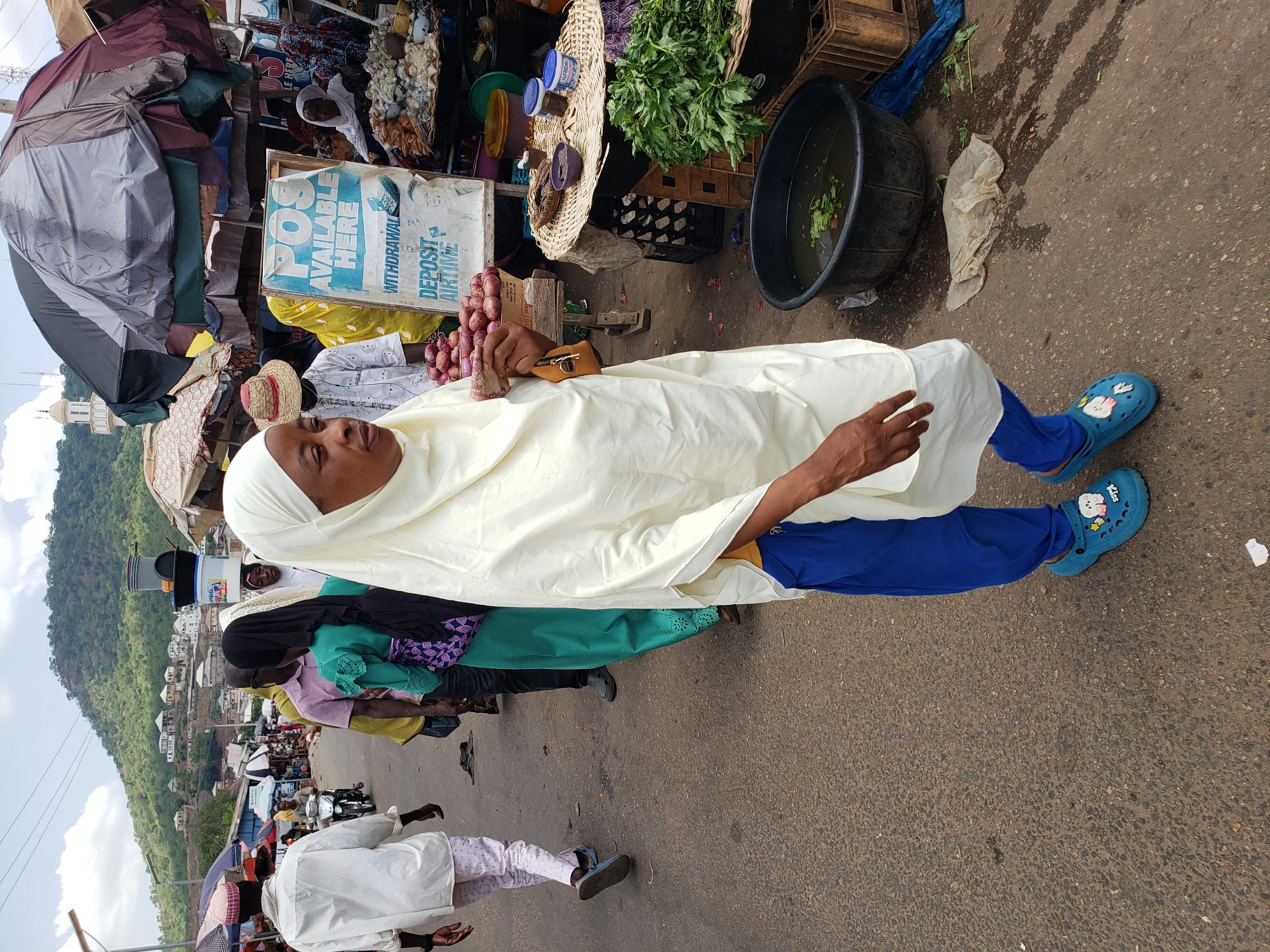 This media file captures a vibrant, everyday scene in an outdoor market in Lokoja, Nigeria. The primary subject is a person dressed in a long, off-white garment (possibly a hijab or similar traditional attire) and blue trousers, wearing light blue clogs adorned with cartoon characters and the word "Kiss." They are holding a bunch of red onions in their hands.
The setting is a bustling street market with various vendors and patrons. In the mid-ground and background, other individuals are visible, mostly women, some wearing headscarves and traditional clothing, engaged in market activities. One person is seen wearing a straw hat. Stalls are set up with large umbrellas providing shade, and goods are displayed in baskets, on the ground, and in crates. Notable market items include leafy green vegetables, piles of red onions, and large black plastic tubs, one of which appears to contain water and greens.
A prominent sign, painted blue and white, reads "POS AVAILABLE HERE" with additional services listed as "WITHDRAWAL," "DEPOSIT," and "AIRTIME," indicating the presence of financial services at the market. Further in the background, a green, hilly landscape with residential buildings is visible under a cloudy sky, suggesting the market is situated near a natural area. The ground is an asphalt surface, showing some wear and scattered debris typical of a busy public space. The time of day appears to be daytime, with ample natural light. The overall atmosphere is one of active commerce and daily life.