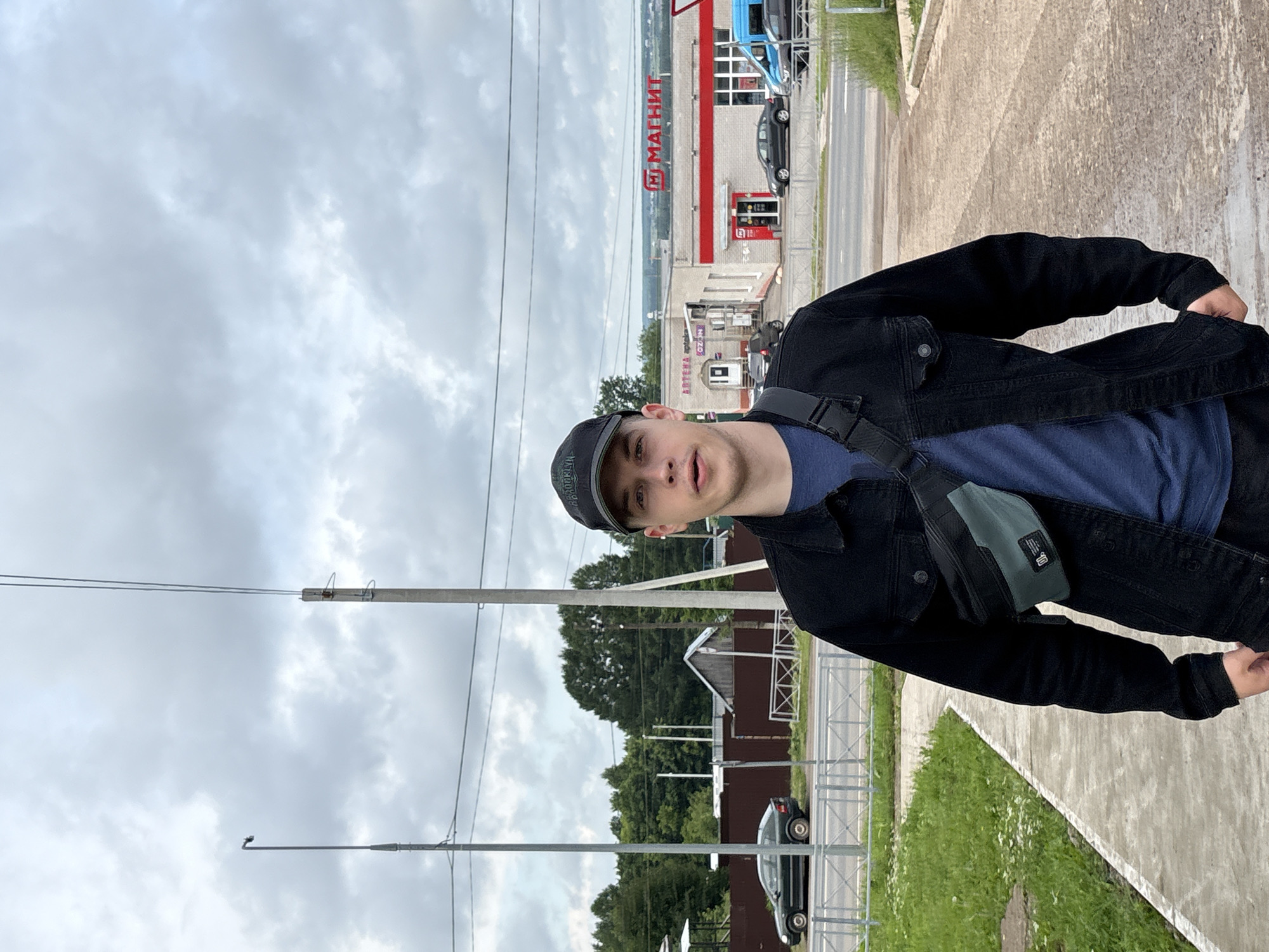A young man stands facing the camera on a concrete sidewalk with a dirt road to his right, in what appears to be a semi-urban or rural area. He wears a dark denim jacket over a blue t-shirt and a black baseball cap. A dark green and black fanny pack is slung across his chest. In the background, across a road and a metal fence, is a row of buildings including a store with a prominent red and white sign that reads "МАГНИТ" (Magnit). There are cars parked and driving on the road, and some trees and houses are visible further in the distance. The sky is overcast. The location is identified as Kstinino, Russia.