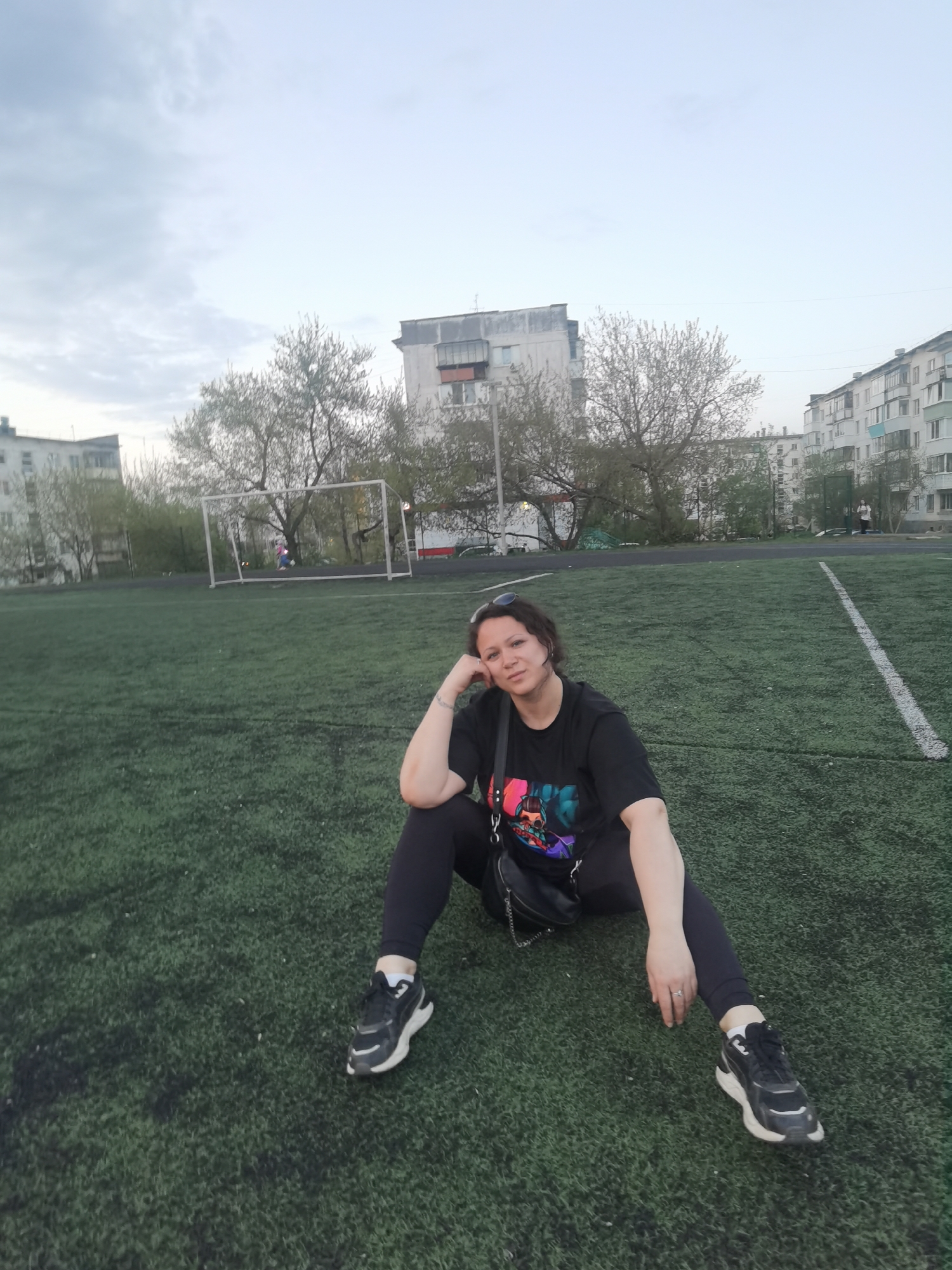 A woman sits on an artificial turf field. She is wearing a black t-shirt, black leggings, and black and white sneakers. She has short, dark hair and is resting her head on her left hand. A black crossbody bag rests next to her. In the background is a soccer goal, a white line on the field, and several apartment buildings. The sky is overcast. This photo was taken in Chaikovsky, Russia.