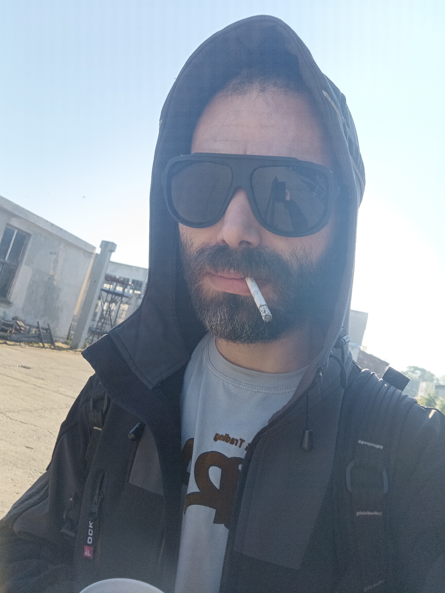 A man with a beard and wearing sunglasses stands outdoors, holding a cigarette in his mouth. He is wearing a hooded jacket and a t-shirt. In the background, there is a building and an open area, possibly a parking lot or industrial area. The sky is clear. Location: Unknown, Unknown.