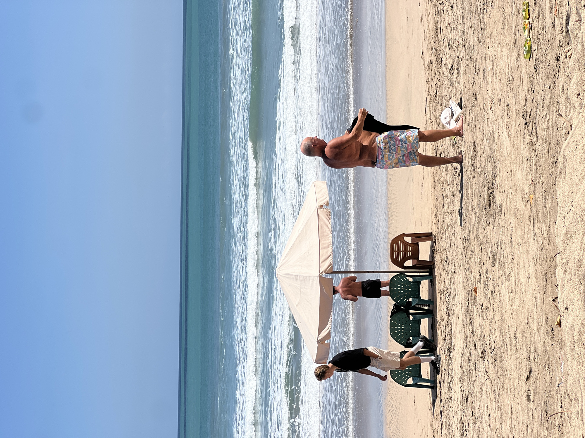 In Kuta, Indonesia, on a sunny day, three men are present on a sandy beach with the ocean in the background. One man, wearing a black short-sleeved shirt and light-colored shorts, walks away from the viewer towards the left. Another man, bare-chested and wearing black shorts, stands under a large, light-colored beach umbrella. The third man, also bare-chested, is to the right of the umbrella, facing left and holding a dark garment. In front of the umbrella, there are four plastic chairs, three of which are dark green and one is brown. The ocean exhibits turquoise waters with white waves approaching the shore.