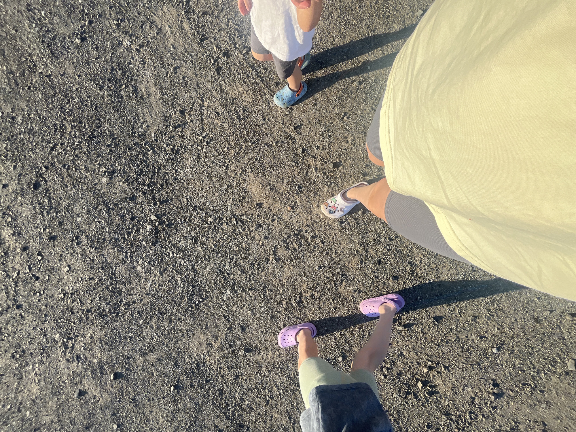 The image shows a ground-level view of several people's feet standing on a gravel surface. Two individuals are wearing purple clogs, one is wearing blue clogs, and another is wearing white clogs with a decorative pattern. One person is wearing gray shorts, another is wearing light green shorts, and a third is wearing dark shorts. A fourth person is wearing dark gray shorts and a light yellow t-shirt. The setting appears to be outdoors, possibly in a park or recreational area, with no discernible urban features or specific landmarks that would indicate a location within Бруснятское, Russia. The lighting suggests it is daytime, with distinct shadows cast on the gravel. The focus is on the footwear and lower legs of the individuals.