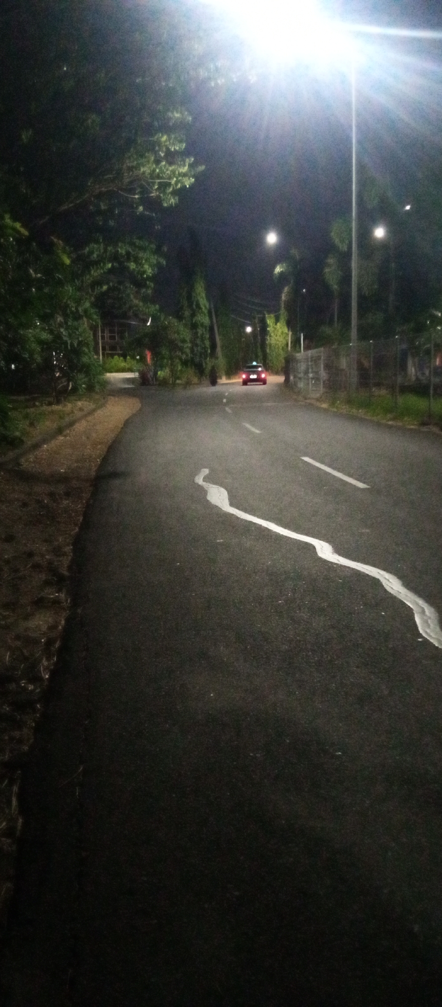 A street scene at night in Surabaya, Indonesia, features a paved road with white lane markings. A red car with its brake lights illuminated is visible in the distance, traveling away from the viewer. The road is lined with trees and vegetation on both sides, with streetlights illuminating the area. A tall lamppost with a bright light source is prominently positioned on the right side of the frame. The ground to the left of the road appears to be unpaved and covered in dirt and grass. A faint white marking snakes across the road surface in the foreground.