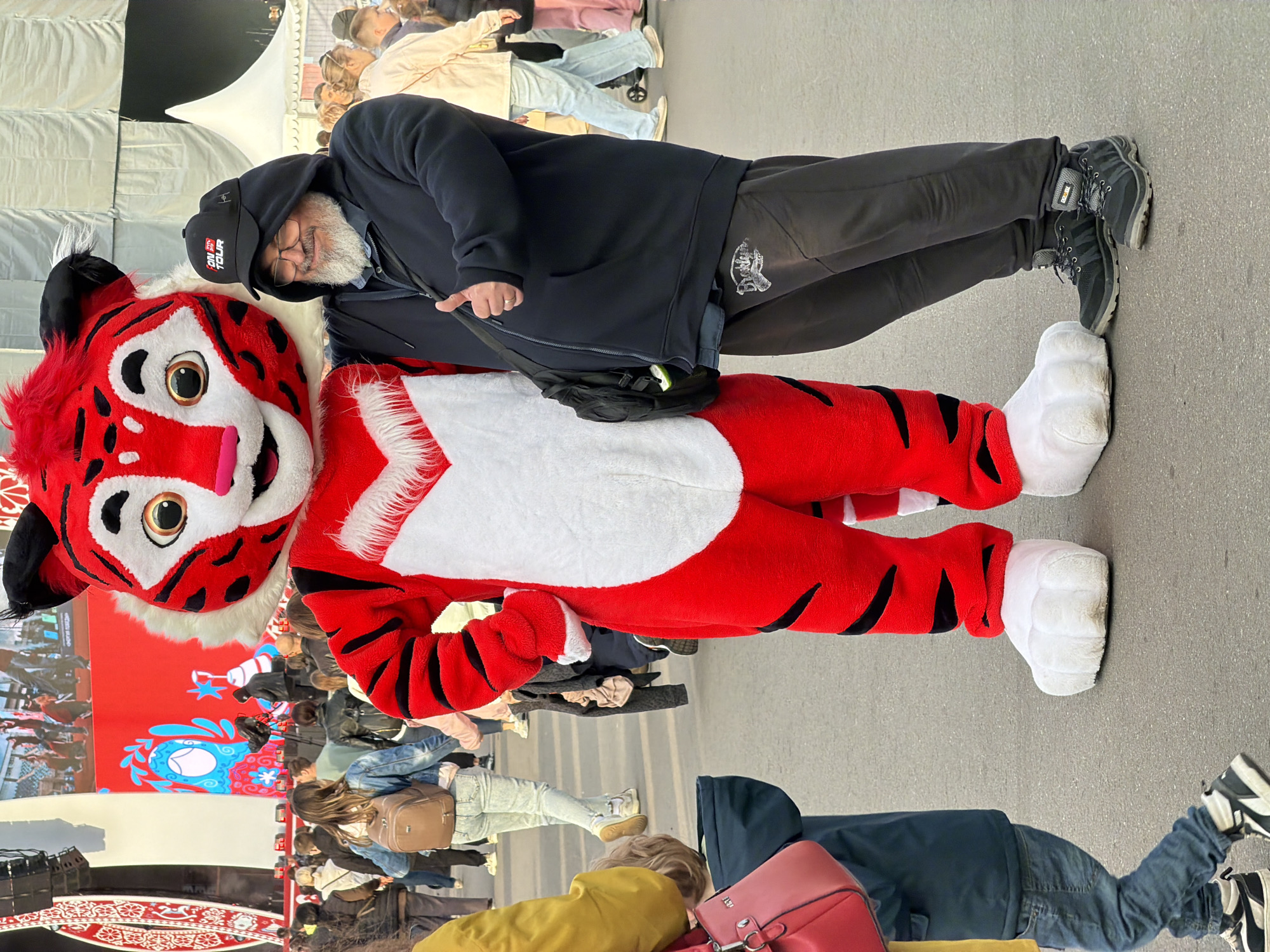 A man in a black hoodie and gray pants stands next to a person in a red and white tiger mascot costume. The man wears a black baseball cap with "ON TOUR" written on it, and he has a gray beard. He is looking at the camera. The mascot is also looking at the camera with a friendly expression. The scene appears to be outdoors in a public area, with a crowd of people in the background. There is a large screen displaying a colorful image behind the mascot. The city is Moscow, Russia.