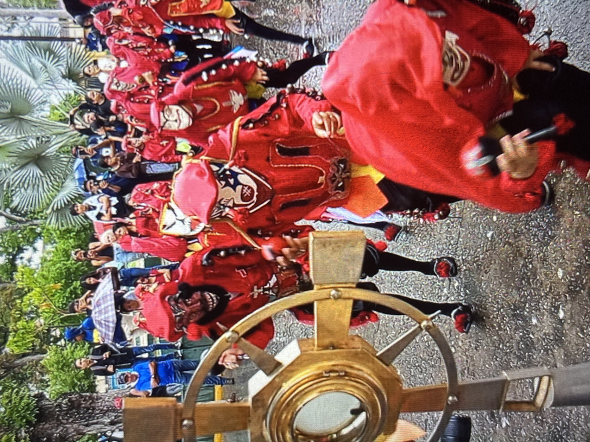 The image depicts a street scene in Tinaquillo, Venezuela, likely during a parade or festival. In the foreground, a person wearing a vibrant red costume and a distinctive mask with black and white facial markings is partially visible, holding a maraca. To the left, a golden, circular object resembling a wheel or astrolabe is prominently featured, mounted on a metal stand. Behind these elements, a group of individuals in similar red costumes and masks are marching, some holding red pom-poms. Further in the background, onlookers dressed in casual attire are observing the procession, with some holding umbrellas, suggesting overcast or rainy weather. Trees and foliage are visible, indicating an outdoor setting within the city.