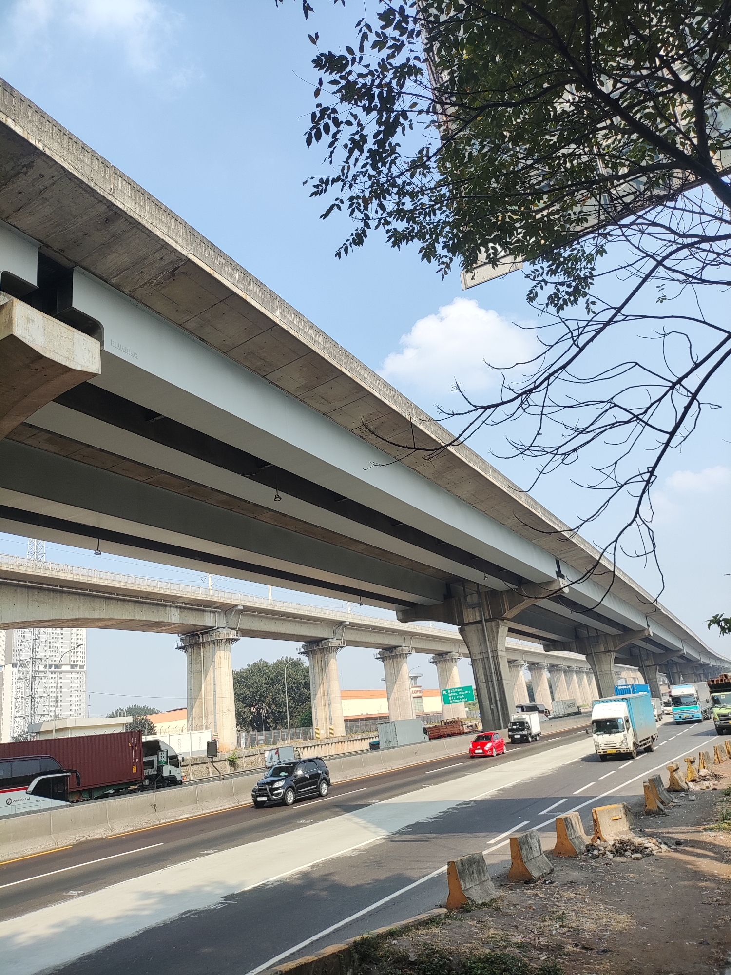 This is a daytime, eye-level, medium-angle, full shot of an elevated highway overpass in Gandamekar, Indonesia. The concrete structure of the overpass dominates the upper portion of the frame, with its massive support pillars extending down to the lower highway level. The sky is a clear blue with scattered white clouds.
Below the overpass, a multi-lane highway is visible, with several vehicles in motion. A white truck with a blue cargo container is in the right lane, followed by a lighter blue truck. To the left of these, a white truck with a red cargo container is also in transit. In the far left lane, a black SUV is traveling. Further left, a white bus with a dark stripe is visible, and behind it, a large red cargo container is being transported by a truck.
On the right side of the highway, a line of concrete barriers and some debris are present. The ground next to the highway is covered in dirt and sparse vegetation. Trees and buildings are visible in the background, suggesting an urban or semi-urban environment. A signpost with Indonesian text is also visible on the left side of the highway.