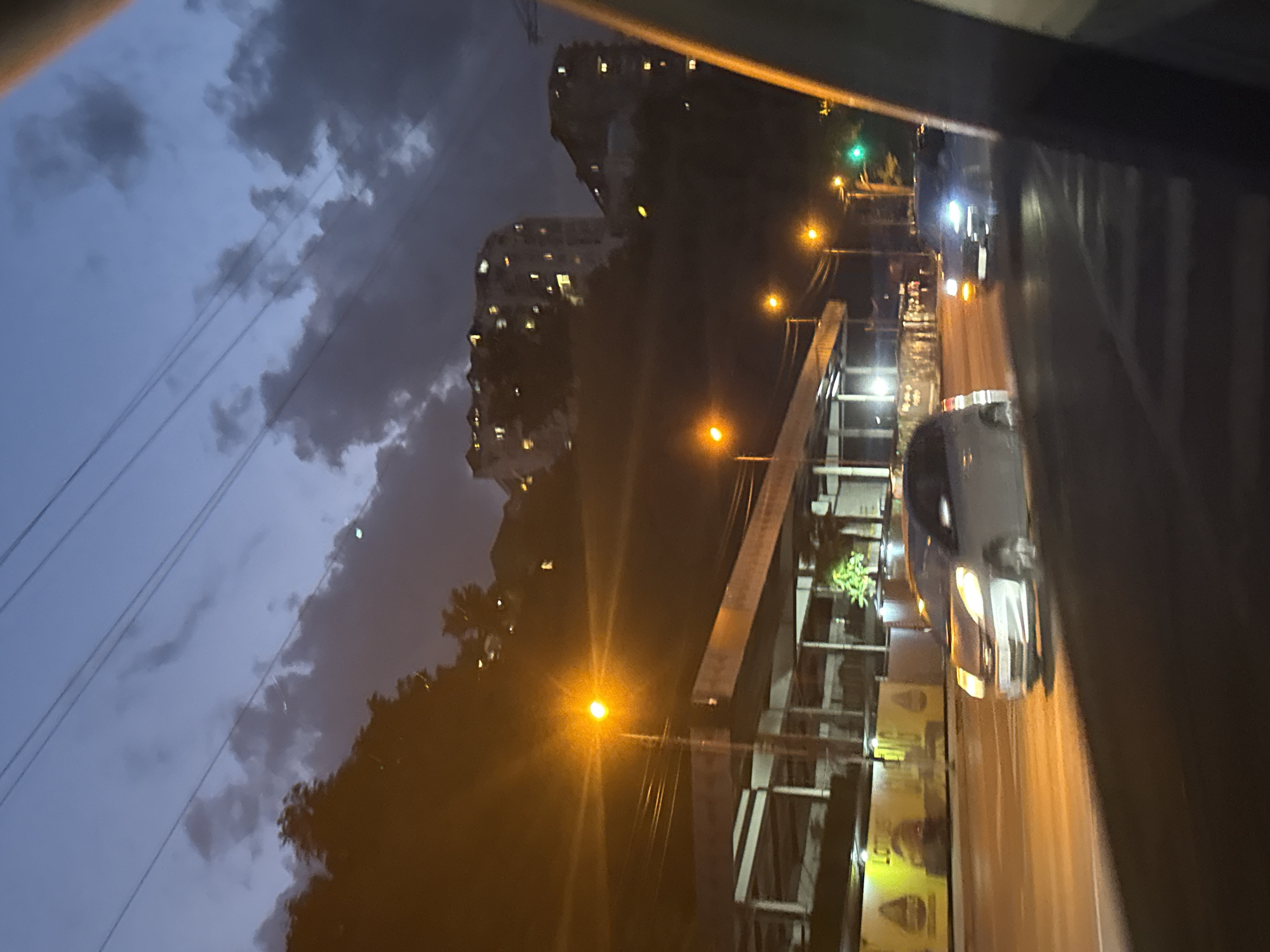The image depicts a twilight scene on a city street, possibly in Tbilisi, Georgia. The sky is a deep blue with scattered dark clouds and faint power lines crisscrossing the frame. In the background, several multi-story buildings with illuminated windows are visible on a hillside. The foreground features a road with moving vehicles. A silver car is seen in motion, its headlights illuminated. Several streetlights cast a warm, orange glow on the surroundings. There are also some trees and what appears to be a structure under construction or renovation along the side of the road. A green traffic signal is visible. The perspective suggests the photo was taken from inside a moving vehicle.