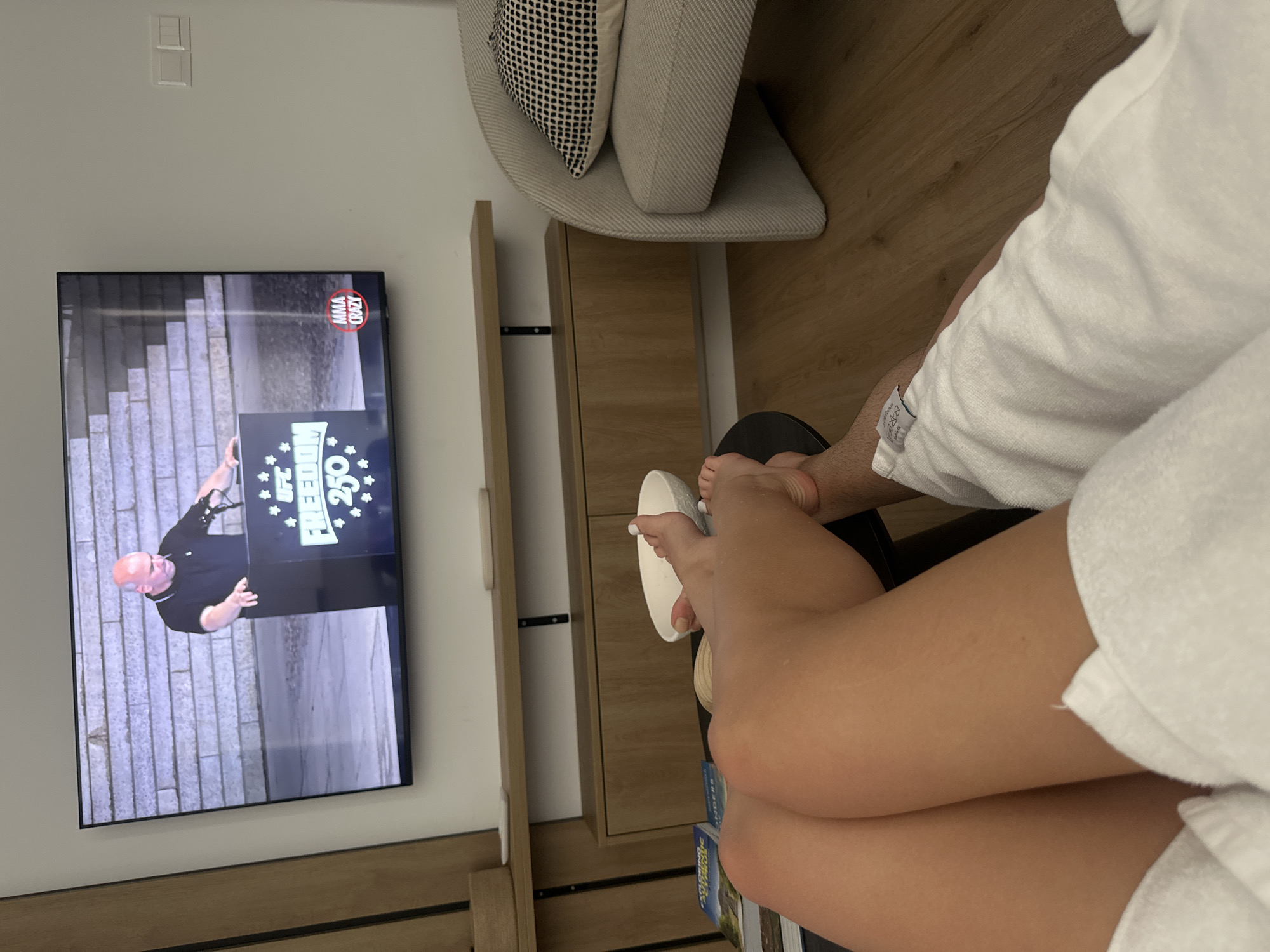 A medium shot shows two people, a man and a woman, relaxing on a floor in front of a television. The man's legs, wearing a white bathrobe, are visible in the foreground, with his feet pointing towards the right. The woman's legs, bare and tanned, are also in the foreground, bent at the knees. They are watching a television screen displaying a man in a black shirt speaking at a podium. The podium has the words "UFC FREEDOM 250" and "MMA CRAZY" on it. The room appears to be a hotel room in Larnaca, Cyprus, with light wood flooring and a minimalist wooden entertainment center. A white armchair with a patterned cushion is visible on the right side of the frame. A small black table with a white bowl and books on it is between the people and the television.