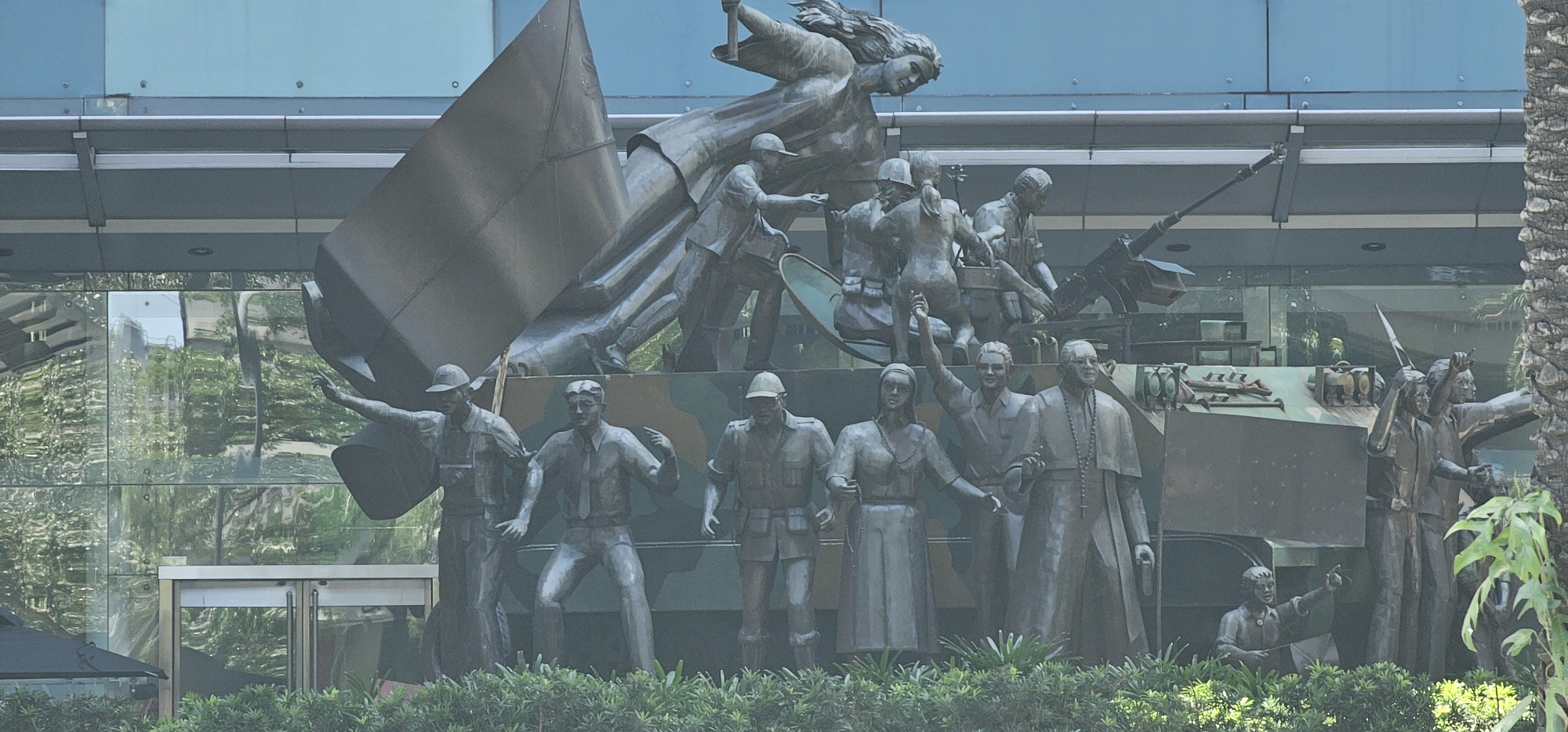 The image captures a bronze monument located in front of the Ayala Triangle Gardens in Makati City, Philippines. The monument, known as the "People Power Monument," depicts a dynamic scene with multiple figures in various poses. At the center, a prominent winged female figure soars above a group of people and a military tank. The figures represent a diverse cross-section of society, including men, women, and a clergyman, some in military attire, others in civilian clothes. They appear to be in motion, with some reaching out, pointing, or holding objects. A large, abstract metallic structure resembling a broken sail or wing is tilted prominently in the background, adding to the sense of drama and movement. The figures are set against the backdrop of a modern building with a glass facade, which reflects the surrounding greenery and sky. The monument is situated amidst lush foliage, with palm trees visible on the right side of the frame. The overall composition conveys a sense of struggle, triumph, and collective action.