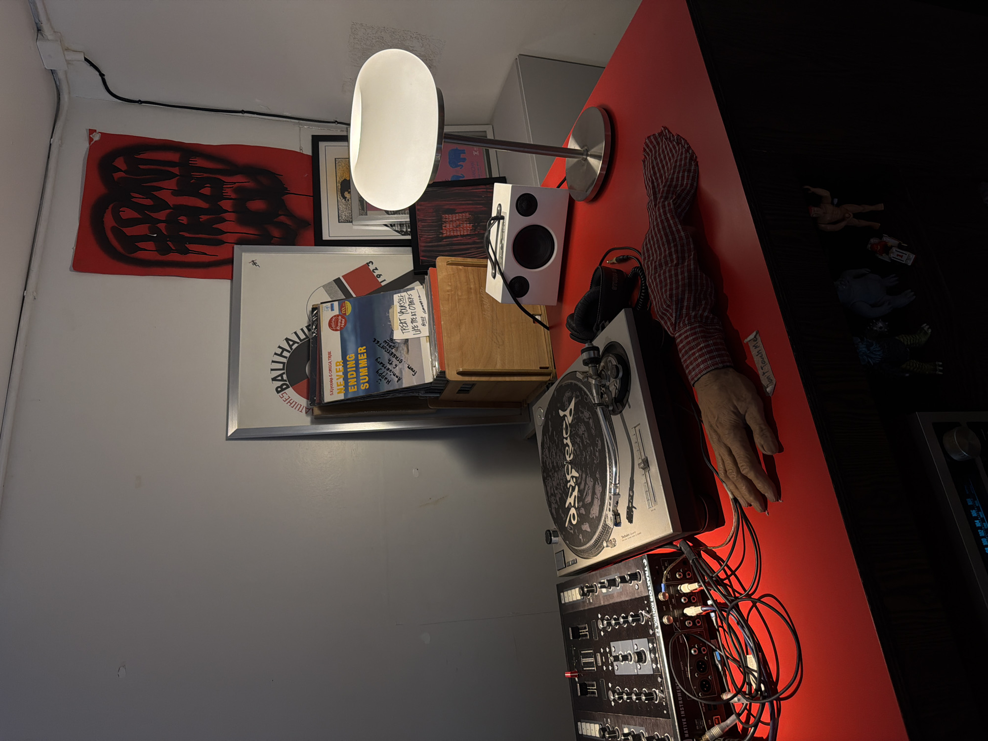 A DJ setup with a turntable and mixer is positioned on a red desk in a room with white walls. A lamp with a white globe shade stands to the right of the turntable. Several records and artwork are displayed on the wall behind the desk. A mannequin arm with a red and white checkered sleeve is placed on the desk, near the turntable. Below the desk, several small toy figures are visible. The overall scene suggests a personal workspace for a DJ or music enthusiast. There is no indication of the specific location within Chiang Mai City Municipality, Thailand.