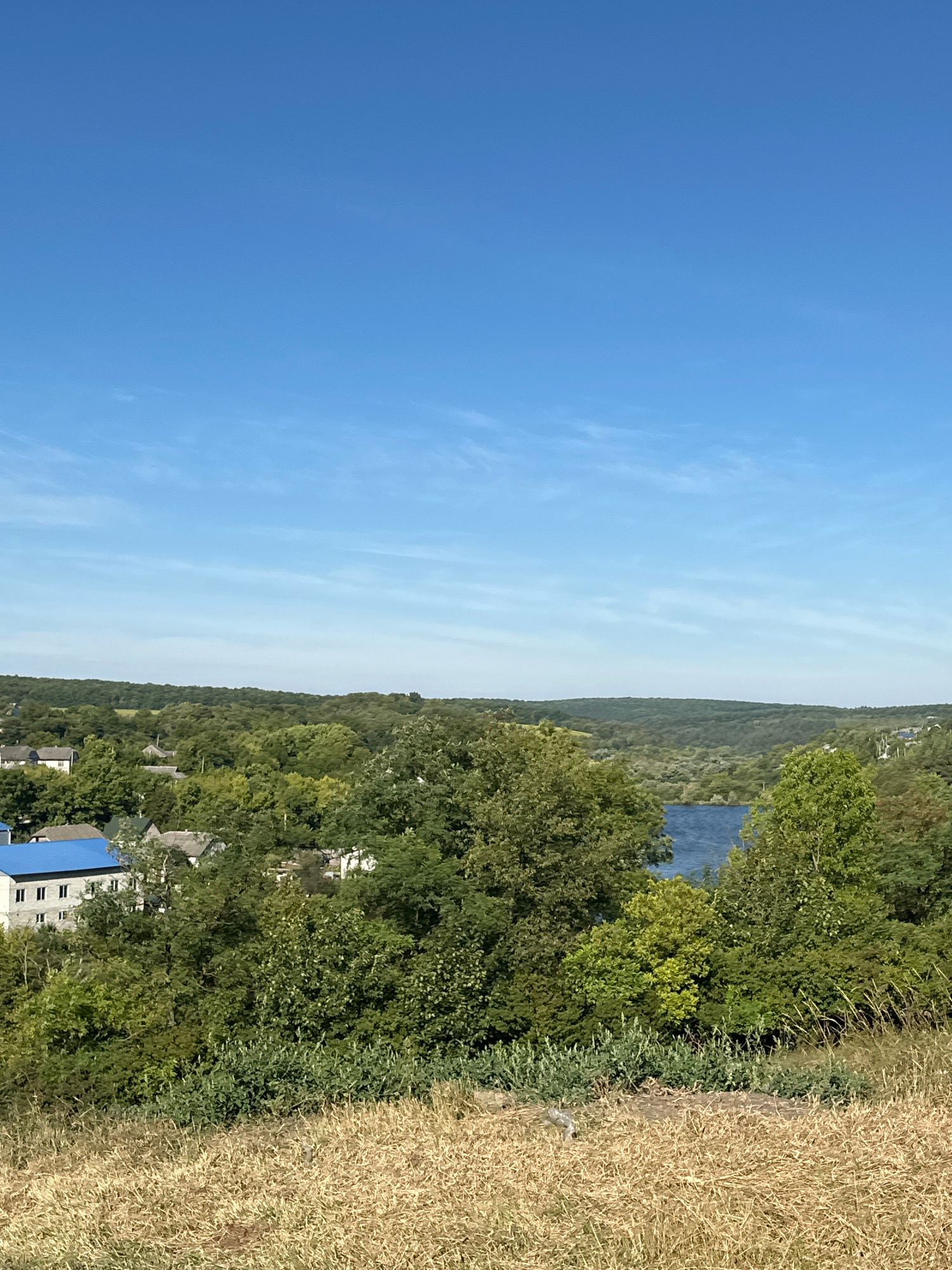 The image shows a landscape view with a clear blue sky and distant rolling hills covered in dense green trees. In the foreground, dry, light-brown grass is visible on a slope. Below the trees, a body of blue water, likely a river or lake, can be seen. A small village or town is nestled among the trees, with several buildings with grey roofs and one building with a distinctive blue roof. The architecture suggests a rural or semi-rural setting. The overall impression is a peaceful, natural environment with human settlement integrated into the landscape. The location is identified as Makiv, Ukraine.