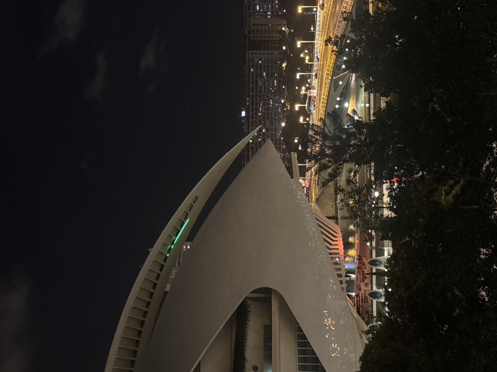 The image shows a nighttime view of the City of Arts and Sciences in Valencia, Spain, focusing on the distinctive architectural elements of the Hemisfèric building. A large, white, curved structure, characteristic of Santiago Calatrava's design, dominates the left side of the frame. This structure has a series of vertical slits along its upper edge, and a bright green light strip runs along its side, illuminating the curved surface.
Below and to the right of the Hemisfèric, a bridge with multiple lanes of traffic is visible, illuminated by numerous streetlights. Cars are driving on the bridge, their headlights and taillights creating streaks of light. In the background, a large apartment building with many illuminated windows stretches across the horizon. In the foreground, dark silhouettes of trees obscure parts of the lower section of the scene. The sky is dark with a few wispy clouds. The overall impression is one of a modern, illuminated urban landscape at night.