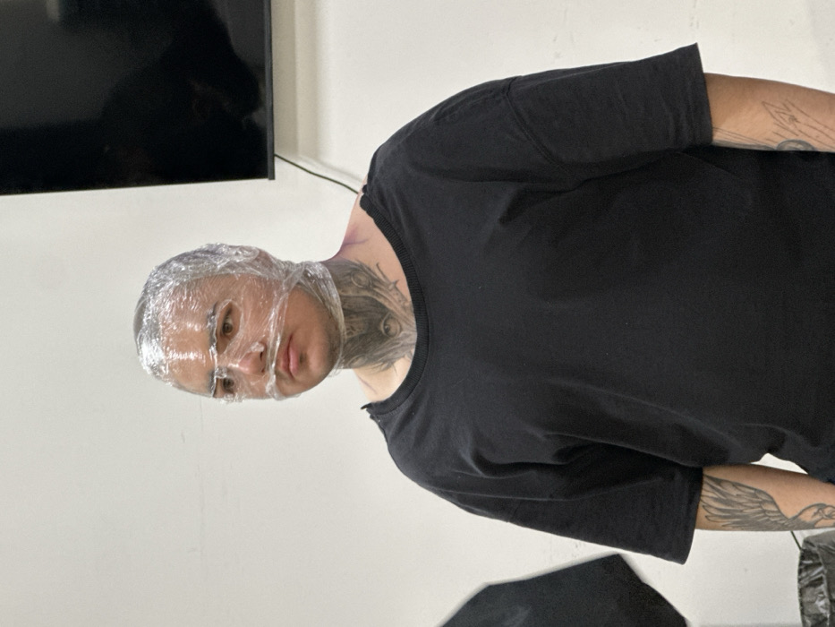 The scene is set indoors in Rostov-on-Don, Russia, against a plain, light-colored wall. A person is depicted standing, facing slightly to the viewer's left, with their head, face, and upper neck tightly enveloped in multiple layers of clear plastic film. The plastic conforms closely to the individual's facial structure, leaving the eyes partially visible through the material, while the nose and mouth are covered. The individual has a neutral expression. Extensive dark ink tattoos are prominent on the visible neck area, featuring an intricate design that includes an eye motif centrally positioned below the chin, with additional patterns extending along the throat and sides of the neck. Further dark ink tattoos are partially visible on both forearms, including a wing-like design on the left forearm. The person wears a loose-fitting, short-sleeved black garment, characterized by a seemingly distressed or cut neckline, particularly on the left side. In the upper right background, a dark, rectangular flat-screen monitor is mounted on the wall. A dark, partially obscured cylindrical object, possibly a waste bin, is visible in the lower-left foreground. The lighting is even throughout the scene.