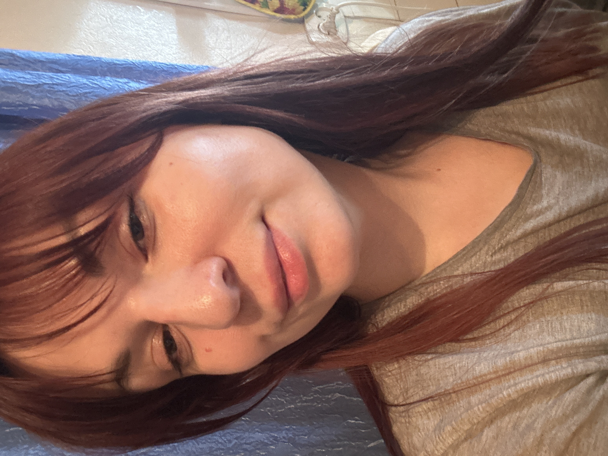 The image depicts a person with red hair lying down. They have fair skin and are wearing a gray shirt. The background includes a blue, possibly textured, surface and a glimpse of other objects out of focus in the upper part of the frame. The image orientation is rotated, suggesting it was taken from a mobile device. There is no information in the image that indicates the location as Фоки, Russia.