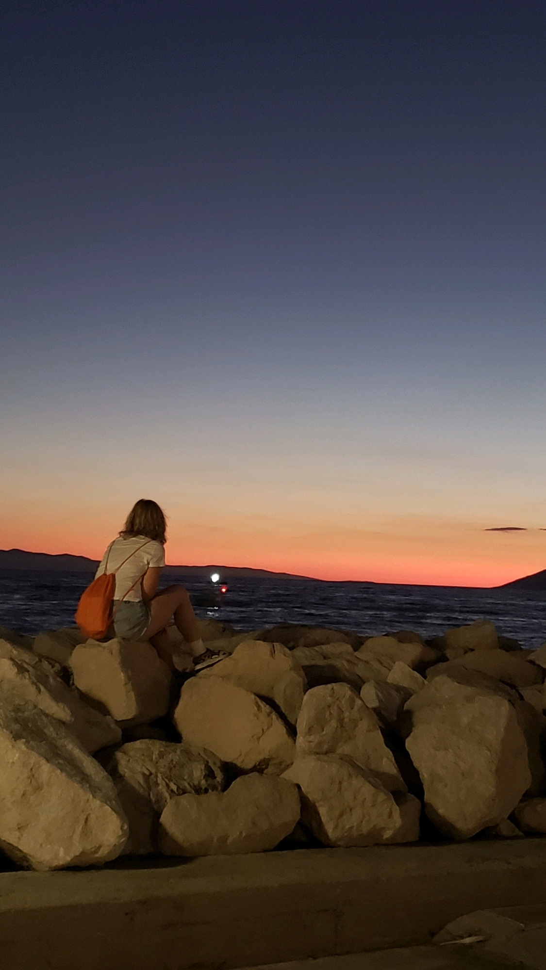 A young person with blonde hair, wearing a white t-shirt and denim shorts, sits on large rocks by the sea at dusk. They have an orange bag slung over their shoulder. The sky transitions from a deep blue at the top to a vibrant orange and pink near the horizon, indicating sunset. The sea is visible, with a faint light or buoys on the water in the distance. Hills or landmasses are silhouetted against the colorful sky. The rocks in the foreground are substantial and uneven, suggesting a natural or man-made breakwater. The scene is likely located in Općina Tučepi, Croatia, a coastal area known for its beaches and scenic views.