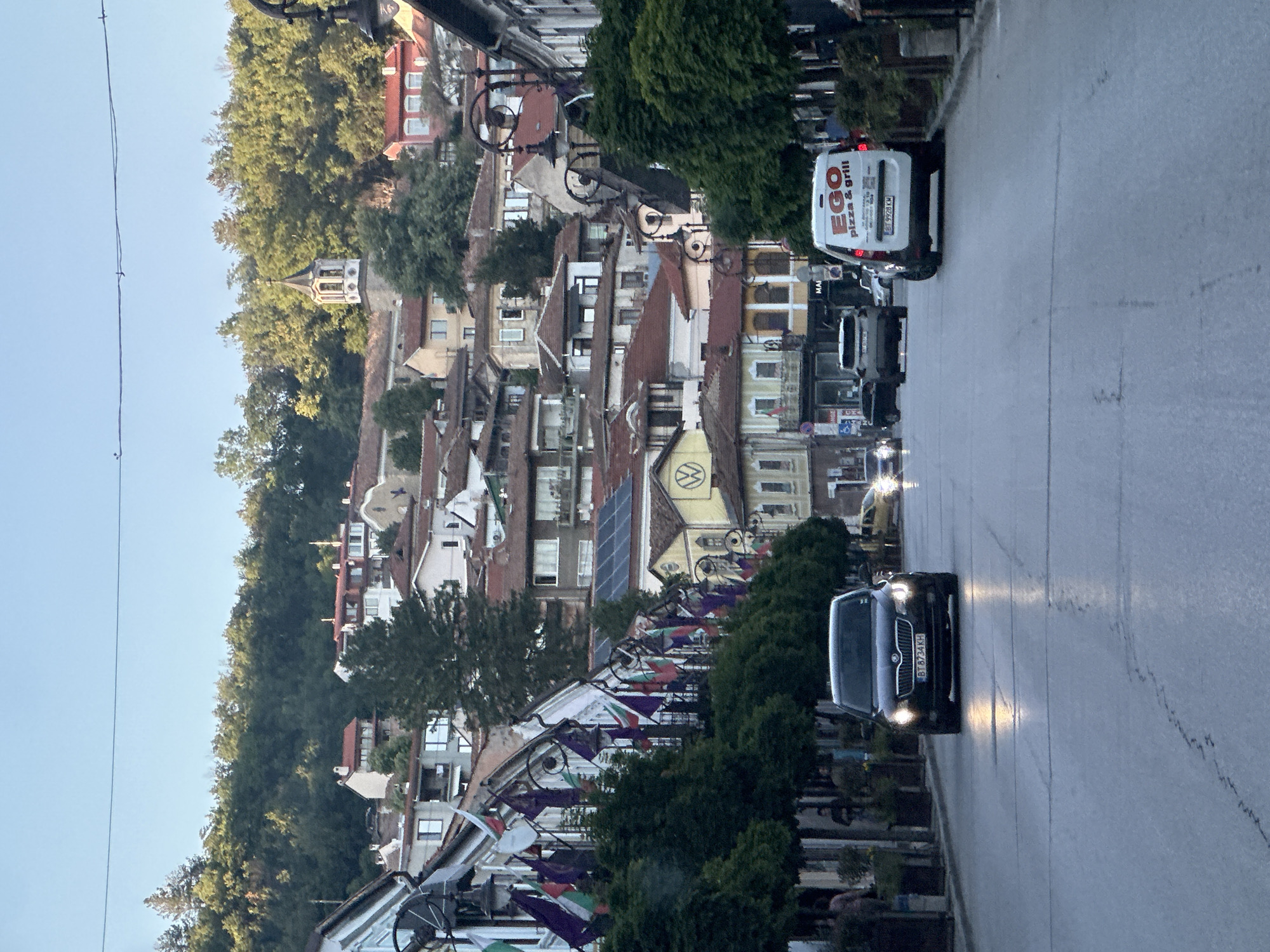 The image depicts a street scene in Veliko Tarnovo, Bulgaria. The street slopes downward, with buildings lining both sides. The architecture is characterized by traditional Bulgarian houses with red-tiled roofs, interspersed with modern structures. The rooftops and facades of the buildings are a mix of red, white, and other colors.
On the right side of the image, several cars are driving on the street. The frontmost car is a dark-colored vehicle with its headlights on, registration BT 6734 KH. Further down, a white van with "EGO pizza & grill" is visible, registration BB 9928 KM. Other vehicles can be seen behind the van.
The left side of the street features trees and decorative elements, including arched structures with bicycle wheels and Bulgarian flags. In the background, a church tower is visible among the trees. The sky is a pale blue.