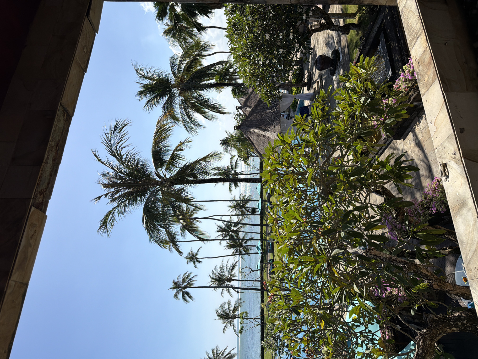 The image depicts a view from a building in Kuta, Indonesia, looking out at a landscape with palm trees and the ocean. The perspective is framed by the architecture of the building on the left and right. In the center, a line of palm trees stretches towards the sea, visible in the background. To the right of the palm trees, there's a garden area with trees, plants, and possibly some structures like a thatched roof covering. The sky is clear and blue.