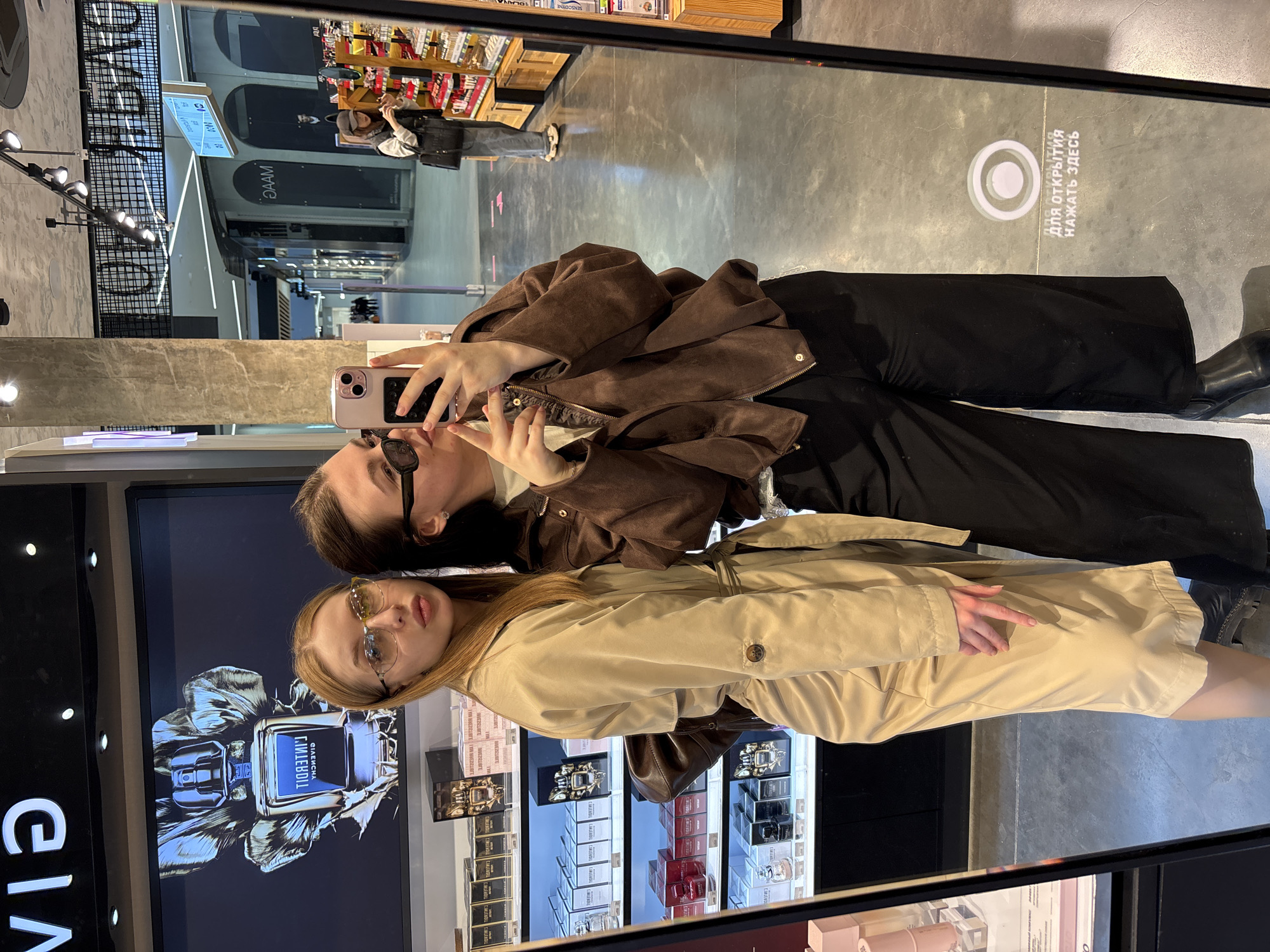 Two young women are captured in a mirror selfie within a retail store, likely a cosmetics or perfume shop, located in Perm, Russia. The woman on the left, with dark hair, wears a brown jacket and black pants, holding a light-colored smartphone to take the photo. She also wears sunglasses. Beside her, the woman on the right, with lighter hair, wears a beige trench coat and glasses, looking towards the camera with a composed expression.
The reflection in the mirror showcases the store's interior, featuring shelves stocked with various packaged cosmetic or perfume products behind the subjects. A large screen prominently displays a reversed image of a perfume bottle, with discernible reversed text "GIVENCHY GENTLEMAN". The floor in the reflection shows a circular graphic with Cyrillic text that reads "ДЛЯ ОТКРЫТИЯ НАЖАТЬ ЗДЕСЬ" (FOR OPENING PRESS HERE), indicating an interactive floor element. The background also reveals reflections of a wider retail environment, possibly a mall corridor, with partially legible and reversed Cyrillic text like "JAAM" and "MAI" on distant signs or walls. The women appear relaxed and are actively posing for the picture during what seems to be a casual shopping trip.