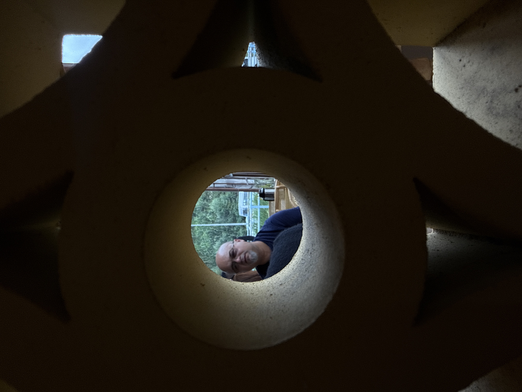 The image captures a man with a bald head and glasses peeking through a circular opening in a textured, light-colored structure. He is wearing a dark blue t-shirt. Behind him, lush green trees are visible, along with a white vehicle and a blue railing. A black thermos and a wicker chair can also be seen in the background. The overall composition suggests an outdoor setting, possibly a park or a cafe in Da Lat, Vietnam, given the prevalent foliage and the casual atmosphere. The foreground is dominated by the dark, abstract shapes of the structure, creating a framing effect for the scene beyond.