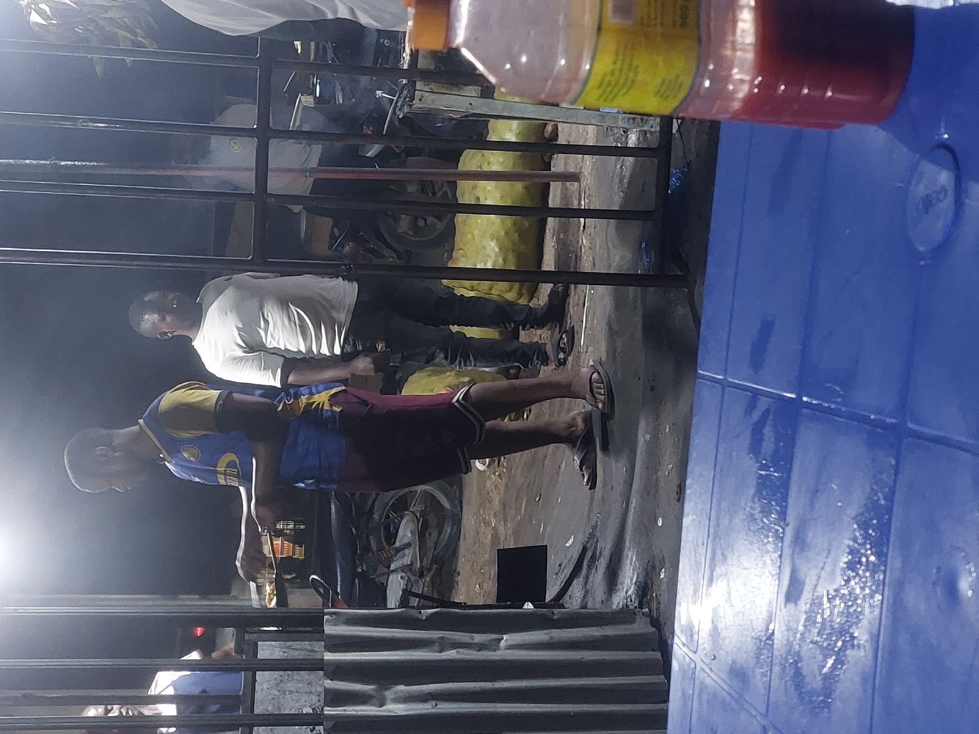 Two men stand in an outdoor setting, likely a street or alleyway, in Dar es-Salaam, Tanzania. One man wears a blue and yellow shirt with maroon shorts and sandals. The other man wears a white shirt and dark pants. They are standing in an area with corrugated metal and metal bars. Behind them, there are motorcycles and other objects, giving the impression of an urban environment. A blue plastic object is visible in the foreground, along with a bottle of liquid.