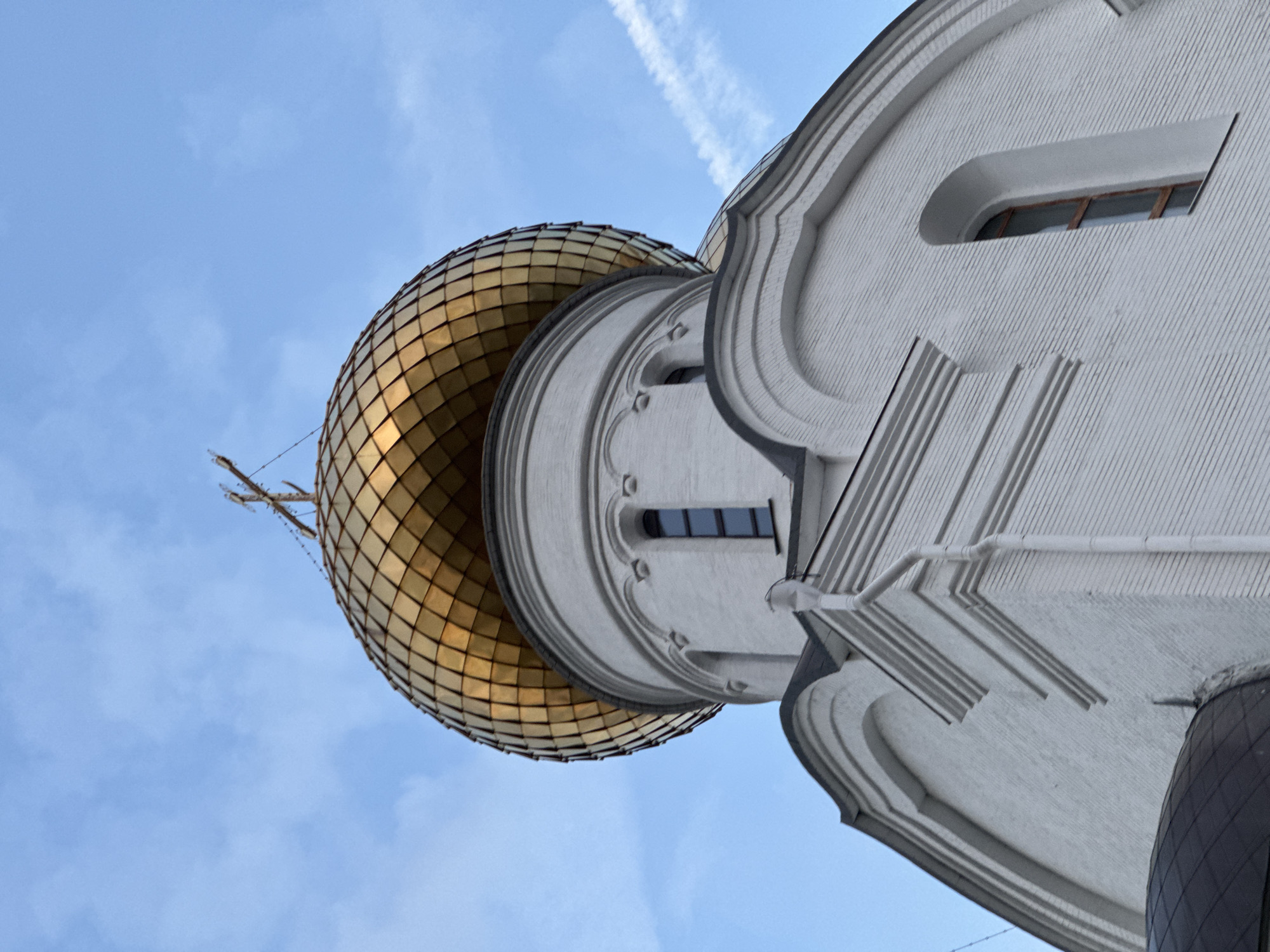 This media file is a close-up, low-angle shot of a white building with a golden dome, set against a blue sky with wispy clouds. The building's architecture is ornate, with decorative moldings and grooved textures on the white facade. A long, thin window with a wooden frame is visible within an arched recess on the building's side. The golden dome is segmented into numerous diamond-shaped panels, giving it a textured, scale-like appearance. The overall impression is one of grandeur and religious architecture.
The image appears to have been taken during the daytime, with soft lighting that suggests it might be morning or late afternoon. There are no people or specific activities depicted; the focus is solely on the architectural elements. The sky is clear but for a few thin, high-altitude clouds, one of which shows a faint contrail, hinting at aerial activity. There are no visible text elements in the image. The orientation of the shot, looking up from a low angle, emphasizes the height and dominance of the structure. The building's design, particularly the golden dome, is characteristic of religious edifices, likely a church or cathedral. The setting appears to be in Шебунино, Russia, as indicated by the location context.