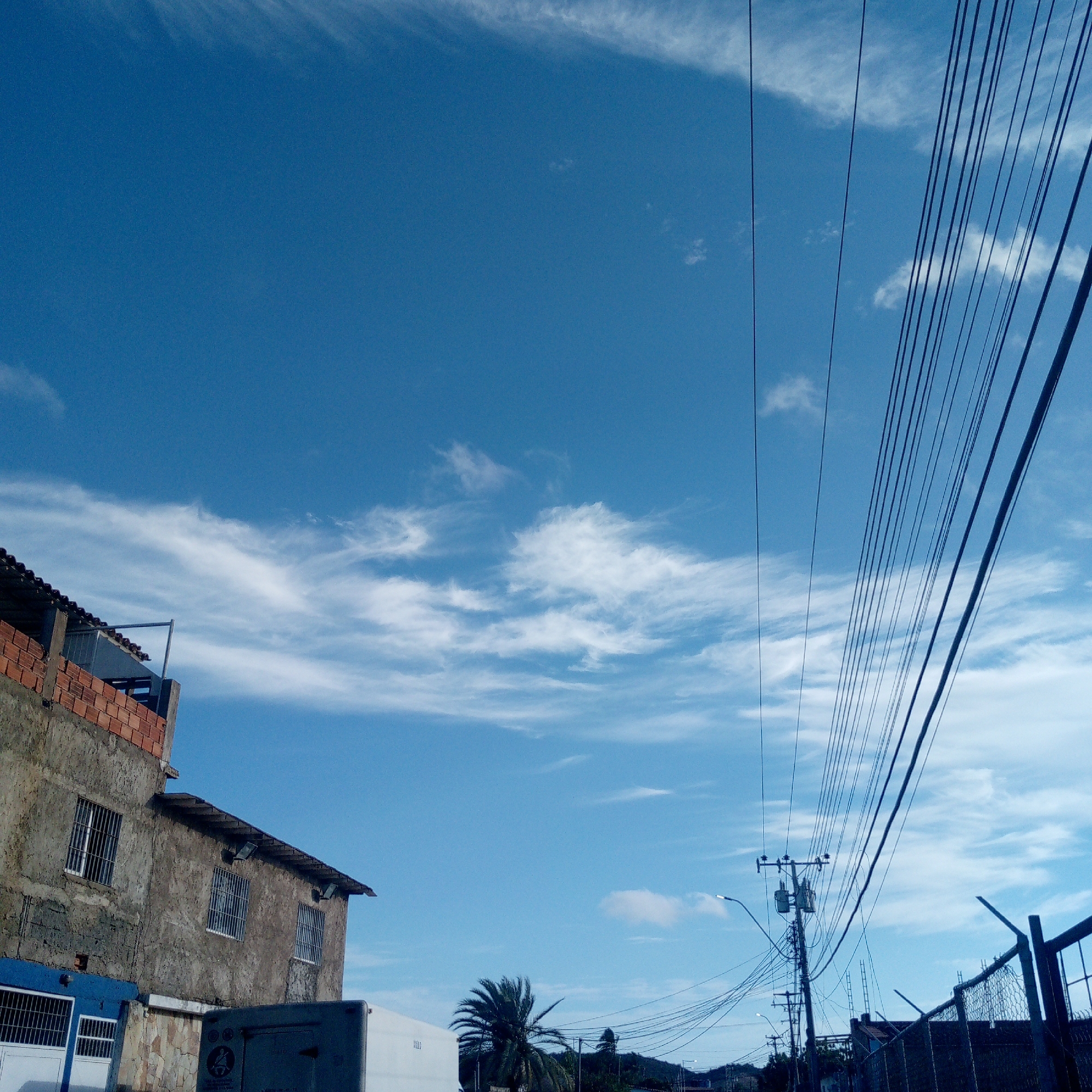 A low-angle shot depicts a street scene in Juan Griego, Venezuela under a partly cloudy, blue sky. Power lines dominate the upper right of the frame, running diagonally from top right to center right. An aged, two-story building with barred windows and a brick extension is visible on the lower left. Adjacent to the building, part of a white truck or van is visible. Further down the street are electrical poles, fences, and a palm tree. A distant mountain range can be seen at the end of the road.
