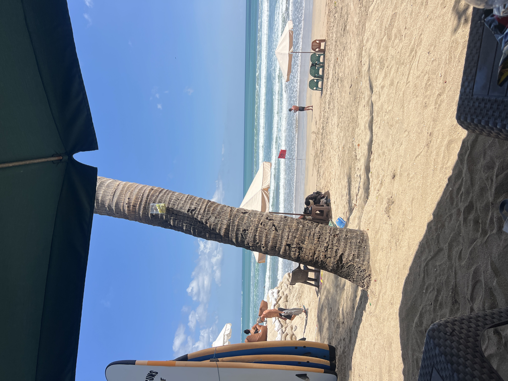 This image depicts a sunny day at Kuta Beach in Bali, Indonesia. The foreground is dominated by a dark, woven patio chair and a table, suggesting a seating area for a beachside establishment. A thick, textured palm tree trunk stands prominently in the mid-ground, casting a dappled shadow on the sandy beach. To the left, several surfboards are propped upright against sandbags.
Further into the scene, a man is walking on the sand, holding a towel. In the distance, the turquoise ocean stretches to the horizon, with gentle waves breaking on the shore. A red flag is planted on the beach, and a few beach chairs are arranged under a light-colored umbrella. The sky is a clear, bright blue with a few wispy clouds. The overall atmosphere is one of a relaxed beach day.