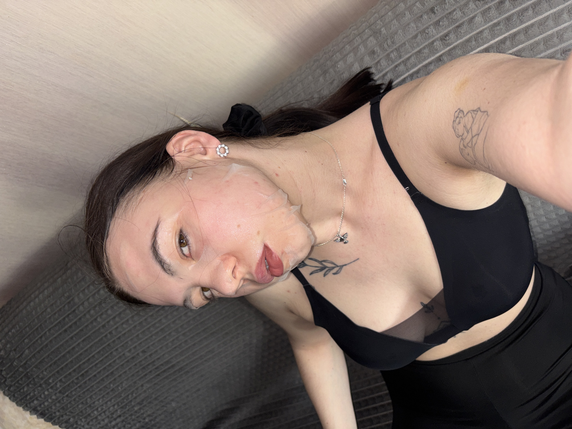 A woman is shown indoors. She is wearing a black bra and dress and has a face mask on. She also has earrings, a necklace, and tattoos. The environment includes a couch. There is no information to identify this location as Perm, Russia.