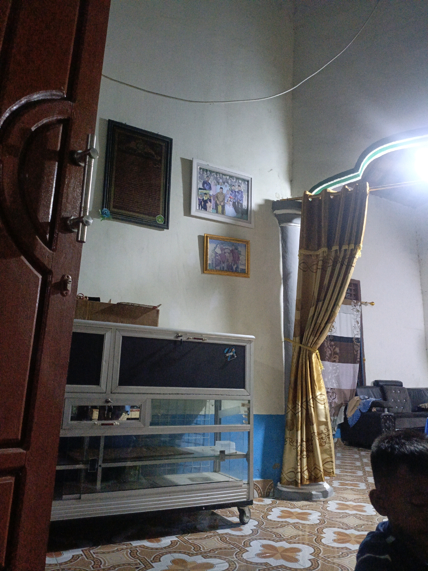 The image depicts an indoor scene, likely a living room or common area in a home in Pagar Ayu, Indonesia. In the foreground, a dark brown wooden door is partially open, revealing its frame and hardware. To the right of the door, a glass-fronted cabinet with metal framing stands on a tiled floor. The cabinet has multiple shelves, some of which are visible through the glass. Above the cabinet, two framed pictures hang on a light-colored wall. The left picture is dark and rectangular, while the right one is smaller and rectangular with a gold frame, displaying a group of people. Further to the right, a thick, golden curtain is tied back and hangs beside a grey pillar. A light fixture illuminates the ceiling, casting a bright glow. In the background, a dark sofa is visible, with some clothing or fabric draped over it. A portion of a person's head and shoulder, seen from behind, is in the lower right corner of the frame. The floor is covered in decorative tiles with a repeating pattern of white, brown, and beige.