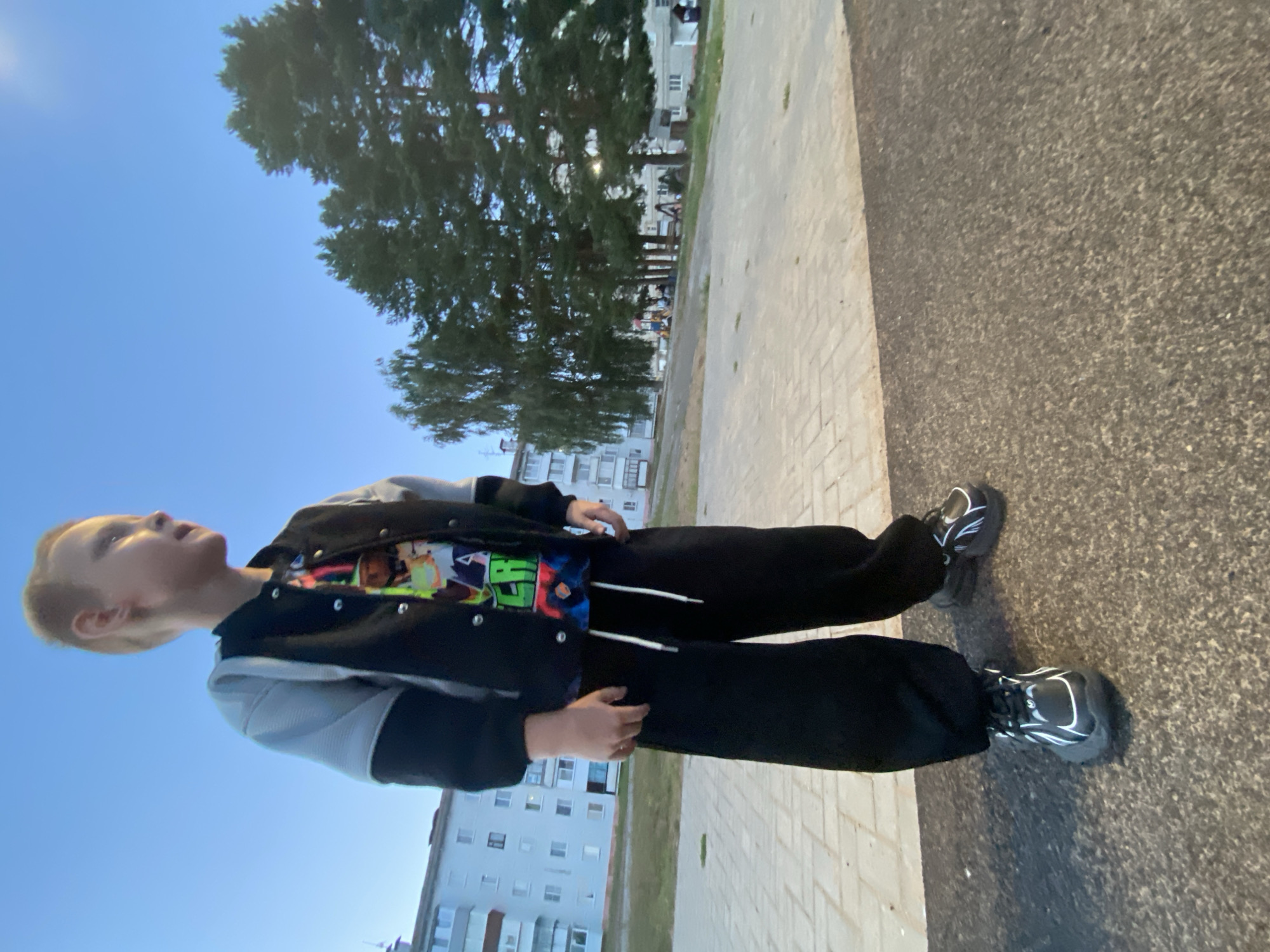 A young boy stands in an outdoor paved area. He is wearing a black jacket with grey sleeves, a colorful t-shirt, black sweatpants, and black sneakers with white accents. He is looking to his right. In the background, there is a multi-story building, trees, and a playground. The sky is clear and blue. The image appears to have been taken in the city of Noviy, Russia.