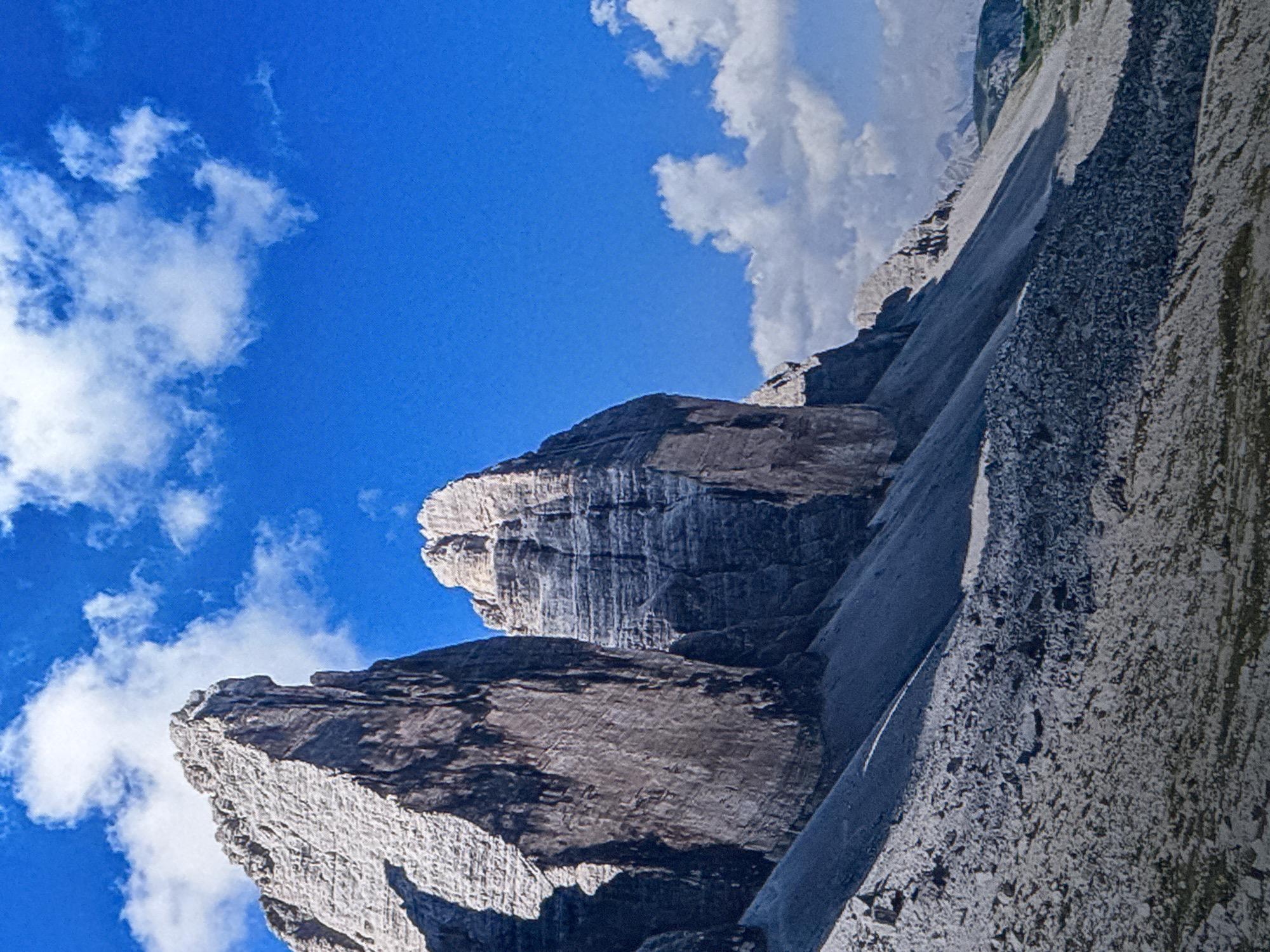 The image depicts the dramatic peaks of the Tre Cime di Lavaredo, a mountain range in the Dolomites, a section of the Southern Limestone Alps in Italy. These iconic formations are characterized by their sheer rock faces, layered stratifications, and jagged summits. The foreground is a scree slope covered in loose rocks and stones, leading up to the base of the mountains. The sky is a vibrant blue, dotted with fluffy white clouds, suggesting a clear and sunny day. The lighting creates strong contrasts, with some rock faces illuminated by sunlight and others cast in deep shadow, highlighting the rugged texture and contours of the landscape. The image focuses entirely on the natural mountain scenery and does not contain any elements related to the city of Saratov, Russia, or any people, objects, actions, or interactions.