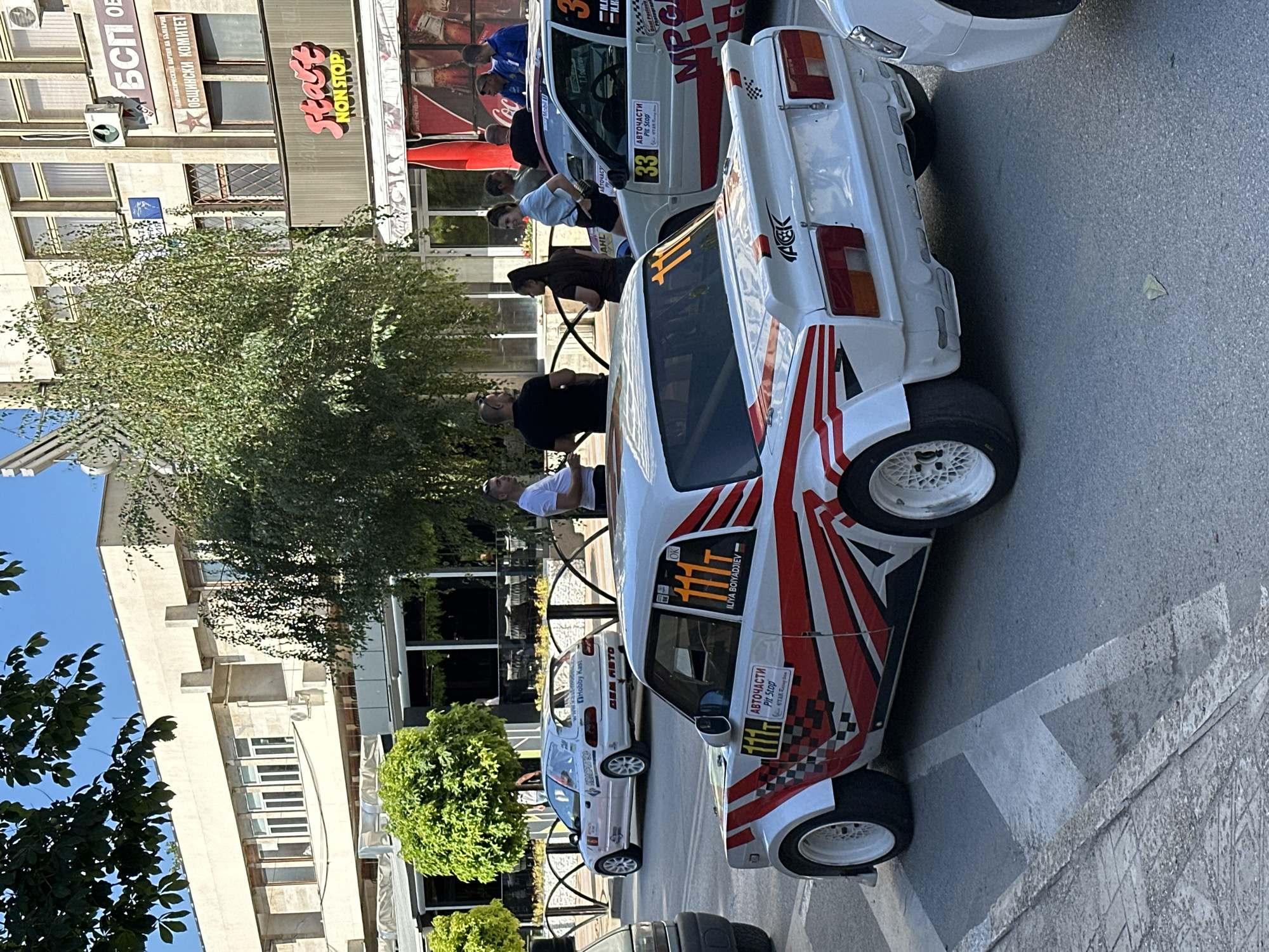 The image depicts a street scene in the city of Gorna Oryahovitsa, Bulgaria, likely during a rally or car event. In the foreground, a white race car with red and white checkered and striped racing decals is parked. The number "111" is prominently displayed in orange on the car's sides and rear. Another white race car, with the number "33" visible, is parked next to it, slightly behind and to the right. Several people are gathered around the cars, some appearing to be participants or spectators. In the background, there are buildings with storefronts, including one with a prominent "Start NON STOP" sign. Trees and foliage are visible, suggesting an outdoor urban setting. The sky is clear and blue, indicating daytime. The street surface appears to be asphalt with painted road markings.