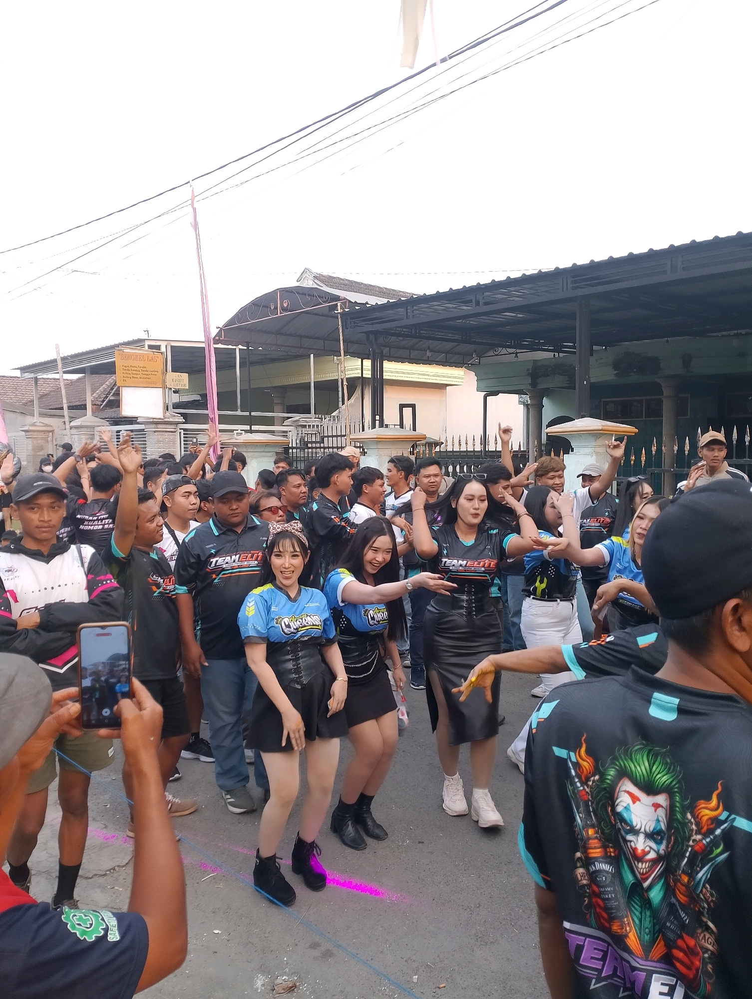 In Rembang, Indonesia, a group of people are gathered outdoors, likely for an event or celebration. The scene appears to be a street, with buildings and a roofed structure in the background. Many individuals are wearing matching black and blue t-shirts with the "TEAM ELITE" logo, suggesting they are part of a team or fan club. Two women in the foreground are prominently featured, dressed in black corsets and short skirts, and appear to be dancing or posing. One of them has a bow in her hair. A man in the foreground is holding up a smartphone to take a picture. Another person in the foreground wears a black t-shirt with a graphic of the Joker and the "TEAM ELITE" logo. The ground is marked with pink powder, possibly indicating a festival or powder-throwing activity. The overall atmosphere seems lively and festive.