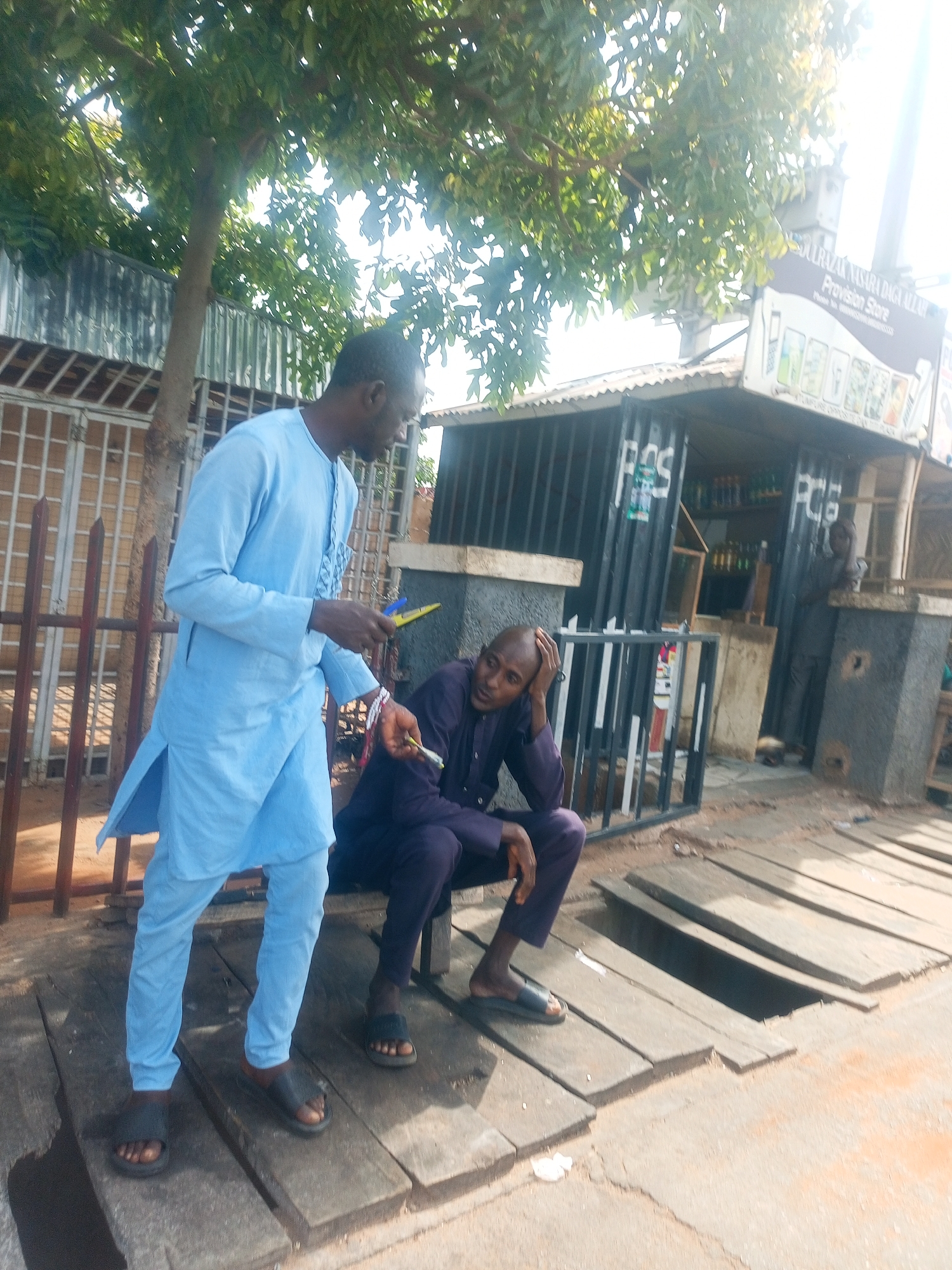 Two men are visible in this outdoor, daytime scene. The man on the left is standing, wearing a light blue traditional African garment with long sleeves and pants. He is holding a yellow object in his right hand and a white object in his left hand. He is also wearing dark-colored sandals. The man on the right is sitting on a wooden structure, wearing a dark purple traditional African garment with long sleeves and pants. He is resting his head on his right hand and wearing dark-colored sandals. Behind the men, a stall with a corrugated metal roof and dark-colored walls is visible. The stall has shelves with bottles and appears to be a provision store, as indicated by a sign that reads "Provision Store" and has images of phones. The environment includes a tree with green foliage overhead, a metal fence, and a wooden walkway constructed from planks. The ground appears to be dirt and sand.