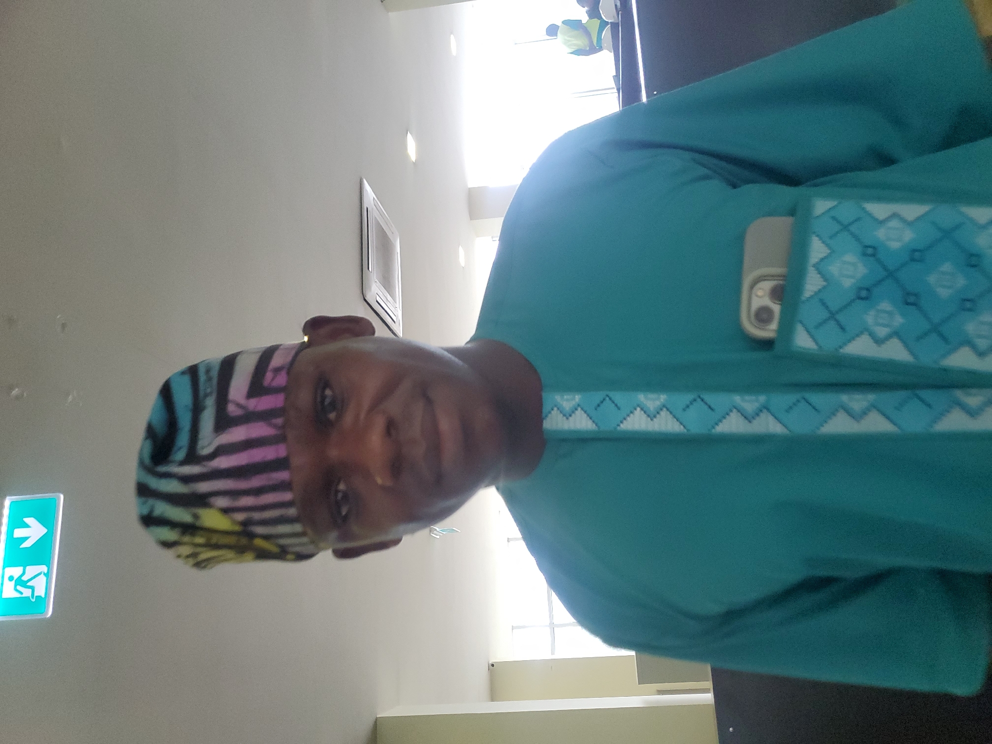 An individual is captured in a close-up, chest-up shot, looking directly at the camera. The person, appearing to be a man, wears a vibrant teal-colored tunic with a patterned trim around the neckline and pocket, featuring geometric designs in white and lighter blue. A smartphone with a multi-lens camera module is visible in the garment's left chest pocket. On his head, he wears a colorful cap or headwear with distinct black, pink, blue, and yellow stripes. His facial expression is neutral, with a slight hint of a smile.
The setting appears to be indoors, likely a well-lit public building or office space. Bright light streams in from the right side of the frame, possibly from large windows or overhead lighting. In the blurred background, several indistinct figures can be seen, with at least one wearing a reflective safety vest. An air conditioning vent or similar unit is mounted on the wall behind the subject. To the left of the frame, a green emergency exit sign with a white arrow pointing right and a running person symbol is visible. The walls are light-colored. The image suggests a casual moment, possibly a selfie or candid photo, taken during the daytime.