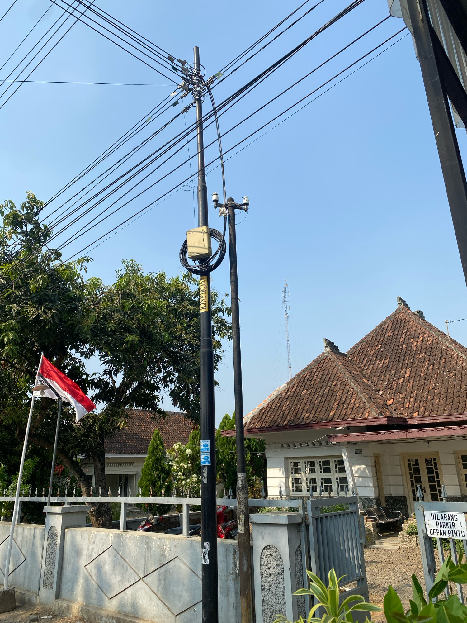 The image depicts a residential street scene in Wonopringgo, Indonesia. A prominent feature is a utility pole with numerous electrical wires crisscrossing overhead against a clear blue sky. Next to this pole stands another, thinner utility pole with some stickers and coiled cables. In the background, a single-story house with a tiled roof is visible, featuring white walls and numerous windows. A fence with decorative patterns runs along the front of the property. To the left, a flag of Indonesia is planted and fluttering. Several trees, including a large leafy one, are present. A distant telecommunications tower can be seen on the horizon. A "No Parking" sign written in Indonesian is placed at the entrance to the property. There are no people or animals visible in the frame. The overall impression is a typical neighborhood street in a sunny climate.