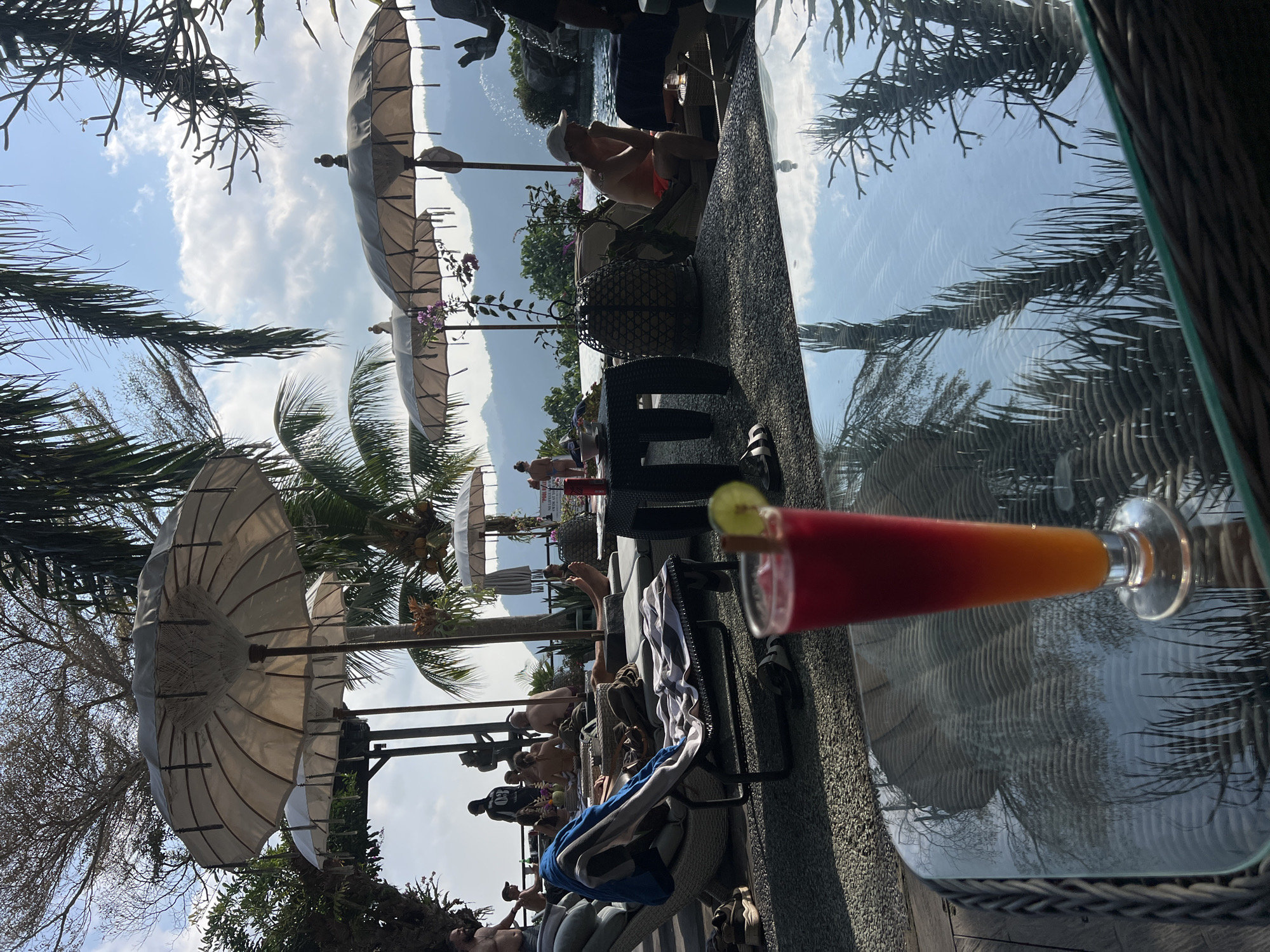 The image depicts an outdoor scene at a resort in Dawan, Indonesia, likely a poolside area with a view of mountains in the background. The foreground features a table with a colorful drink in a tall glass, garnished with a lime wedge and a cinnamon stick. Reflections of palm trees and the sky are visible on the table's glass surface.
In the midground, several lounge chairs are occupied by people relaxing. Some are sunbathing, while others are seated. Umbrellas, typical of tropical resorts, are positioned to provide shade. A small black table with a bottle and a bowl is also visible. A woven basket and more lounge furniture are scattered throughout the area.
The background reveals a swimming pool, a waterfall feature, and lush greenery. Beyond that, rolling hills and mountains are silhouetted against a bright sky with scattered clouds. The overall atmosphere suggests a leisurely vacation setting.